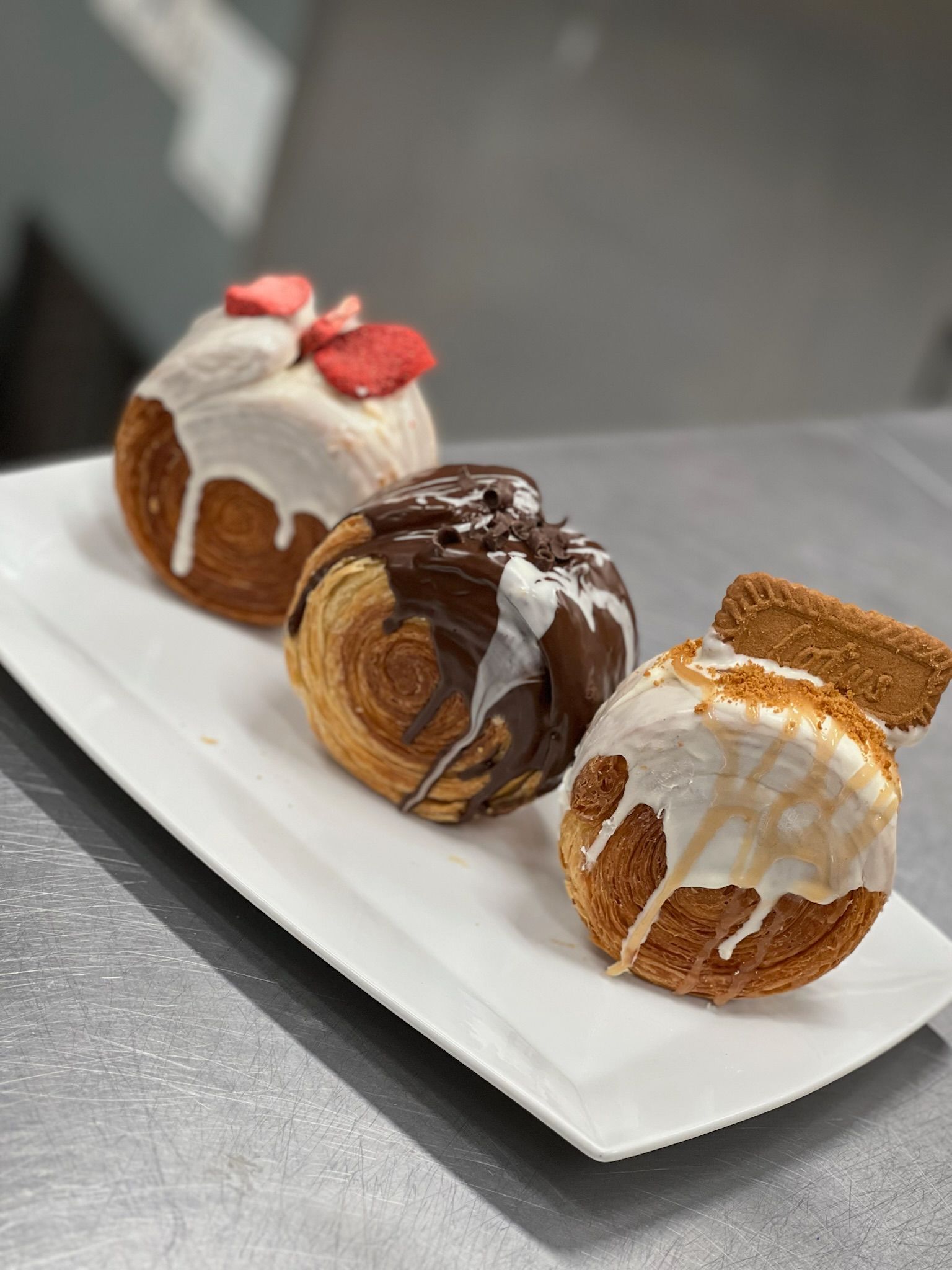 Three croissant-doughnut pastries on a white platter, each with a different topping: strawberries, chocolate, and a cookie.
