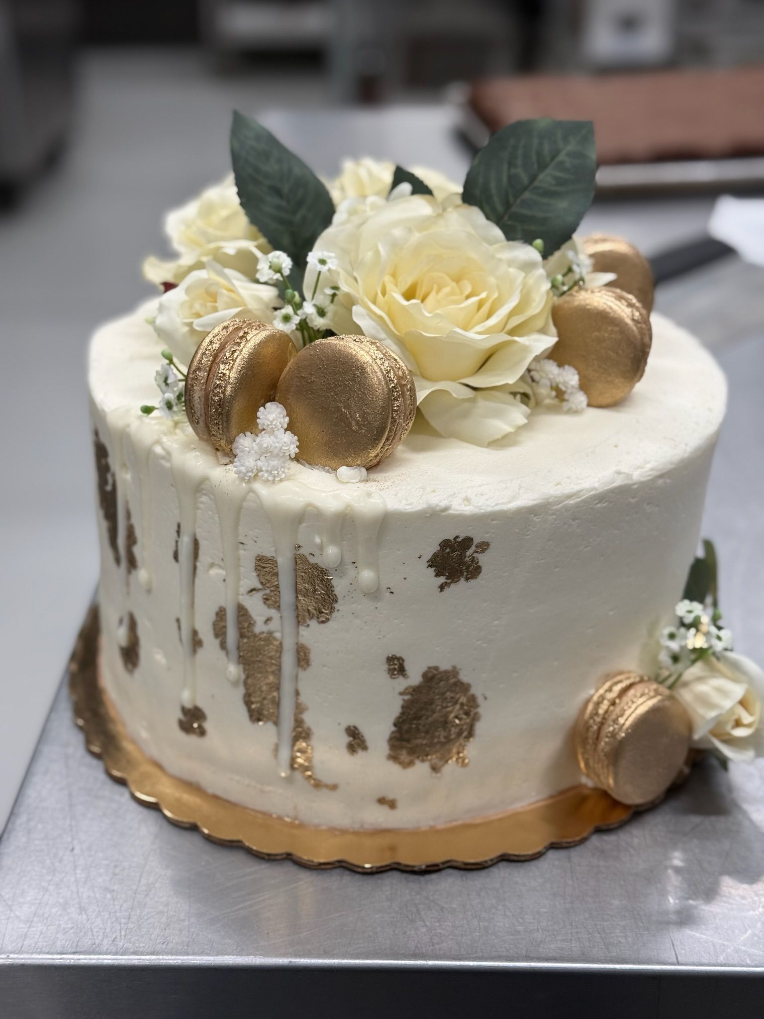 White cake with gold accents, flowers, and macarons on a gold-edged base.