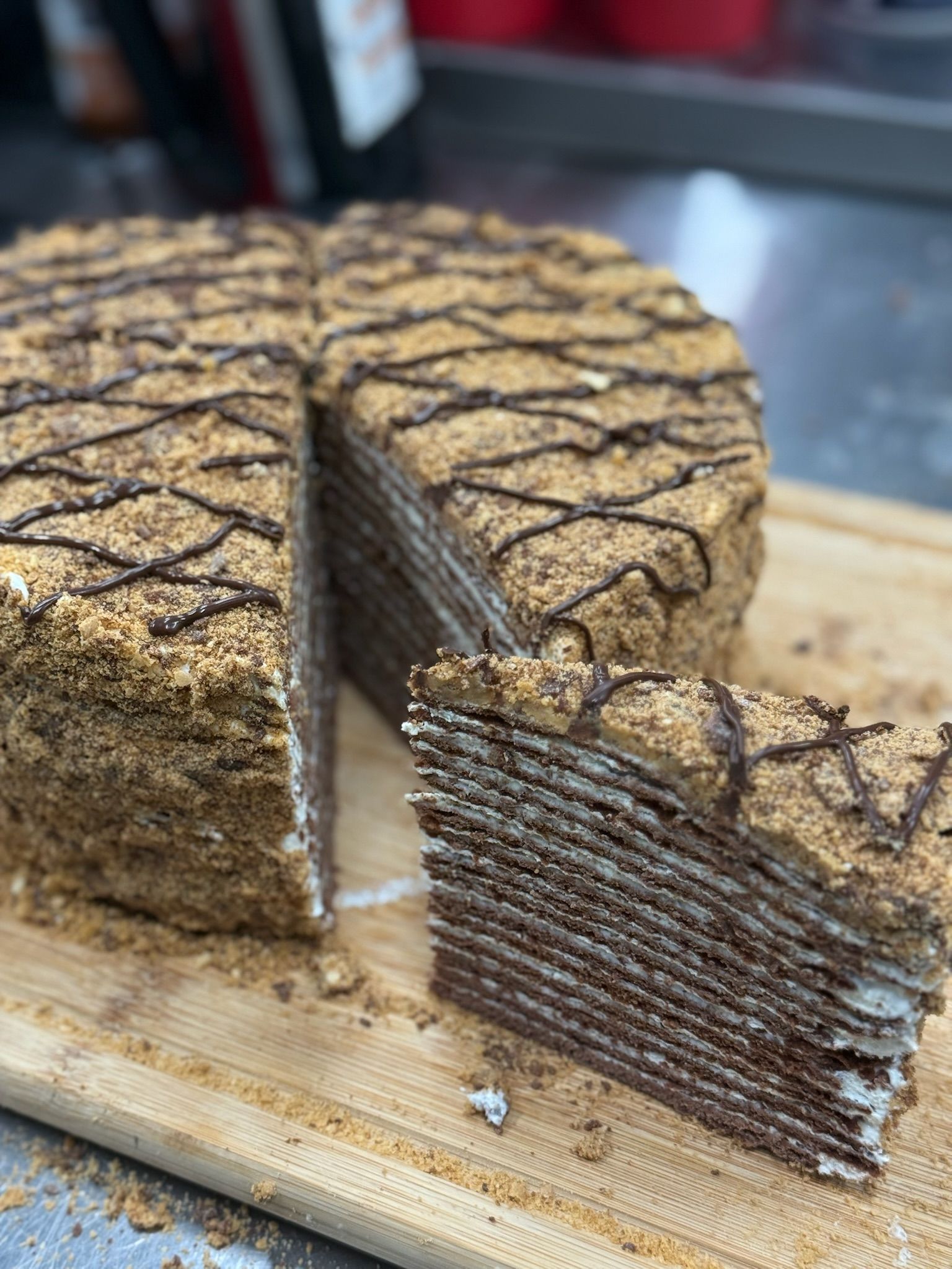 Layered honey cake with a slice removed, drizzled with chocolate and garnished with crumbs, on a wooden board.