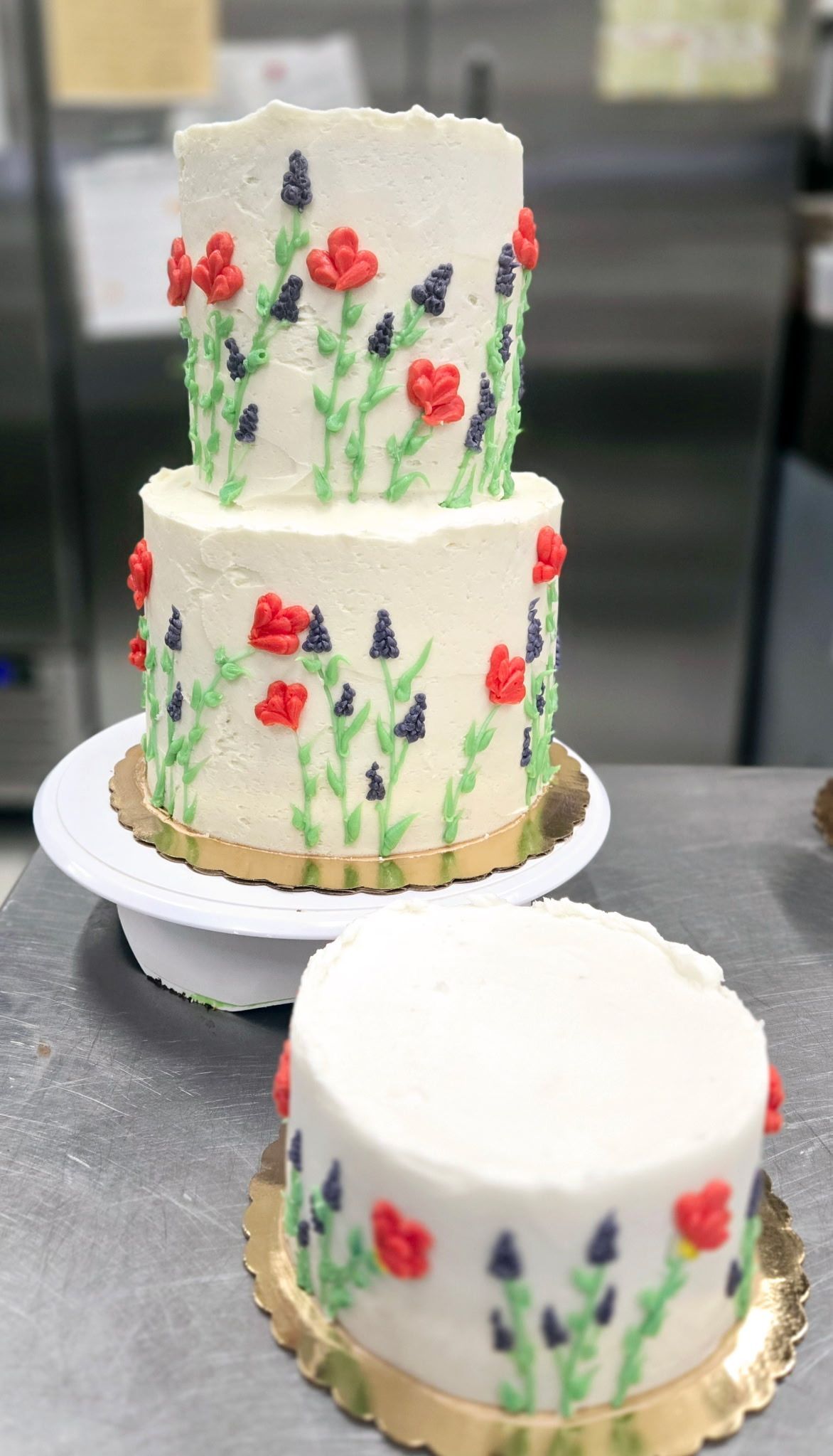 Three-tiered white cake with red and purple flower decorations, on a cake stand and a small separate cake.