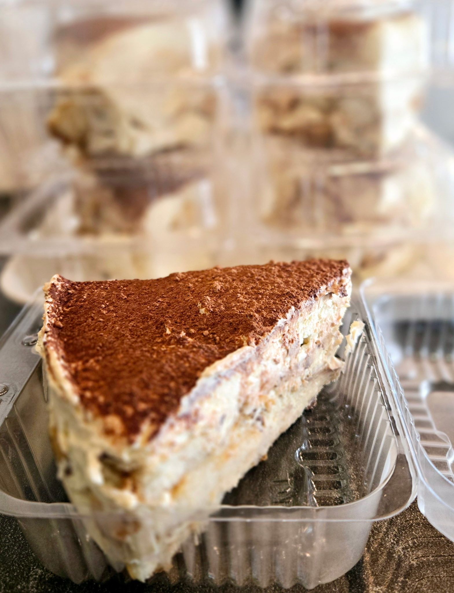 Slice of tiramisu in a clear plastic container, with more tiramisu containers stacked in the background.