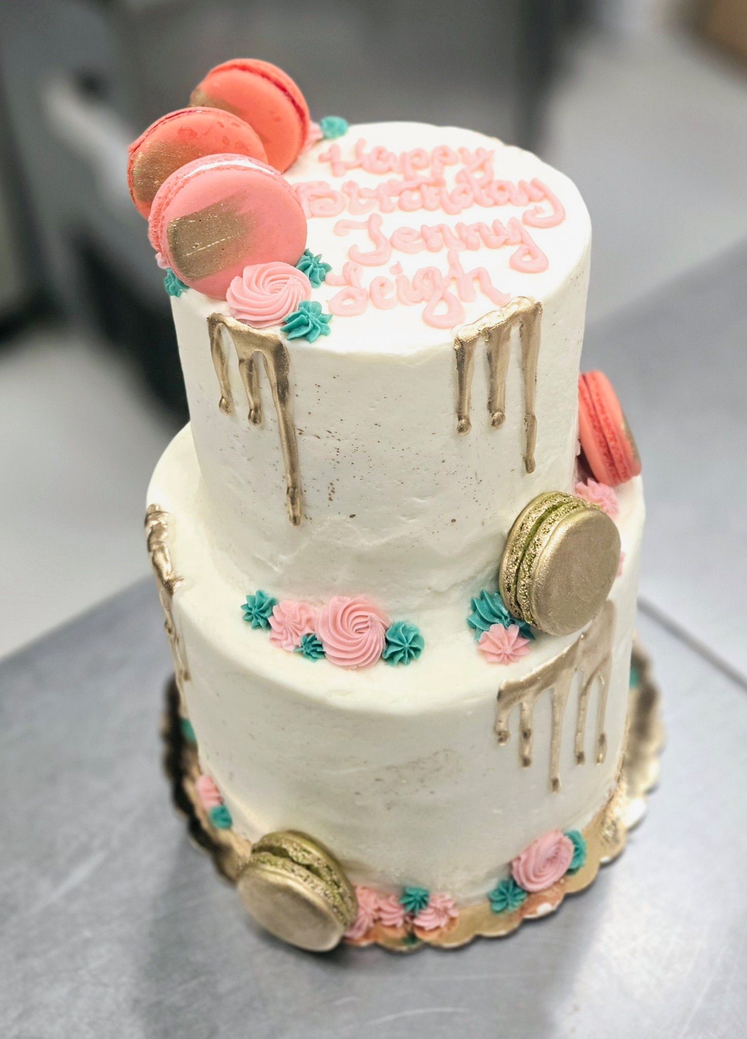 Two-tiered cake with gold drip, pink macarons, and floral frosting.
