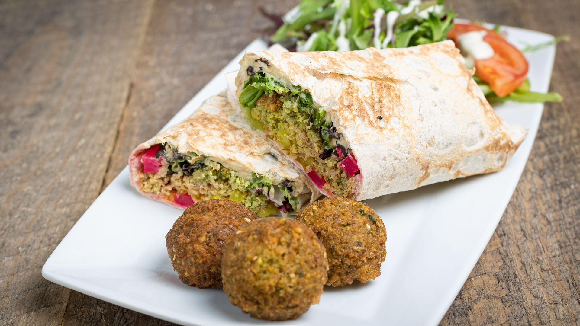 Falafel wrap and balls on a white plate with salad on a wooden table.