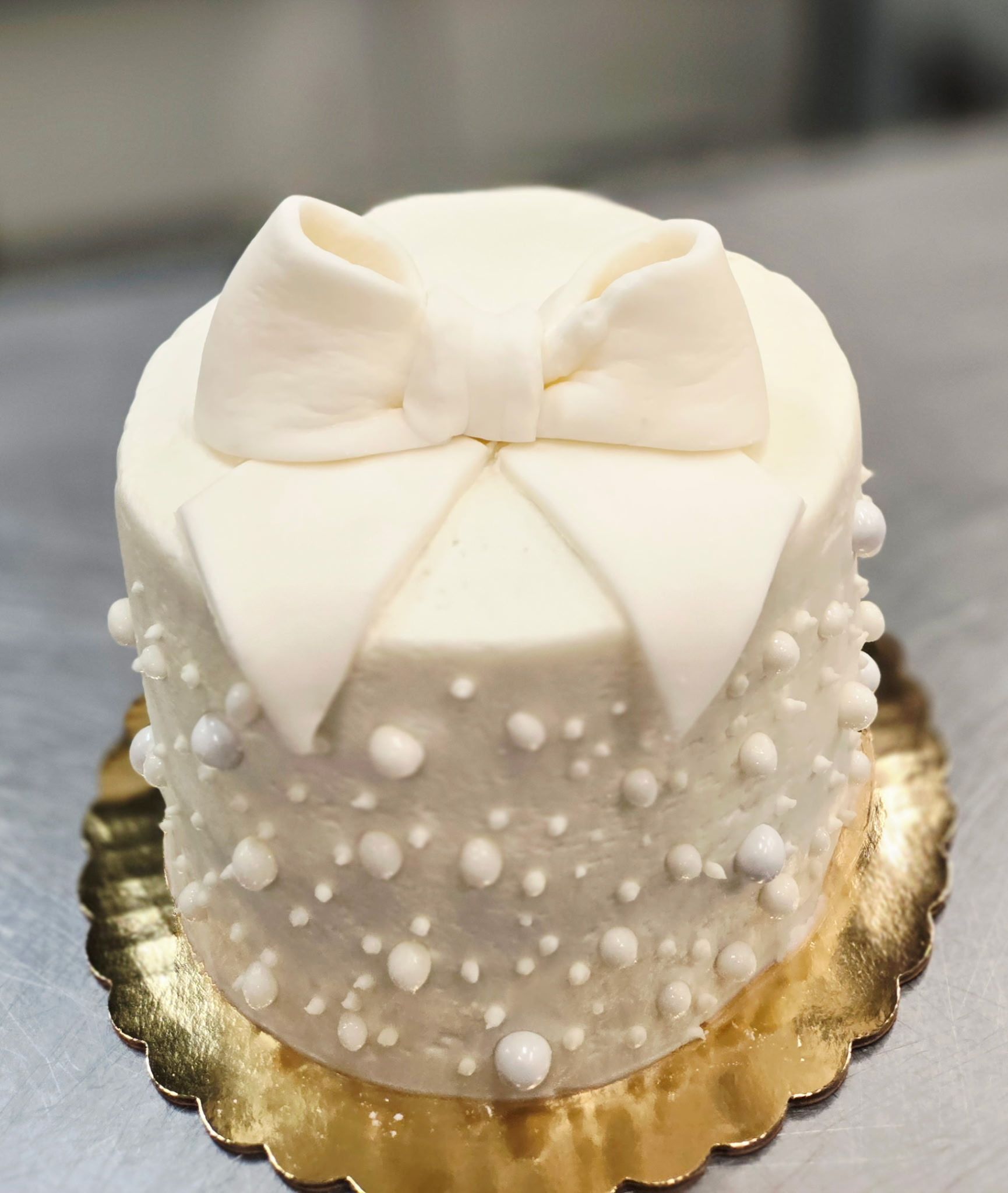 White cake with pearl-like dots and a fondant bow on a gold scalloped base.