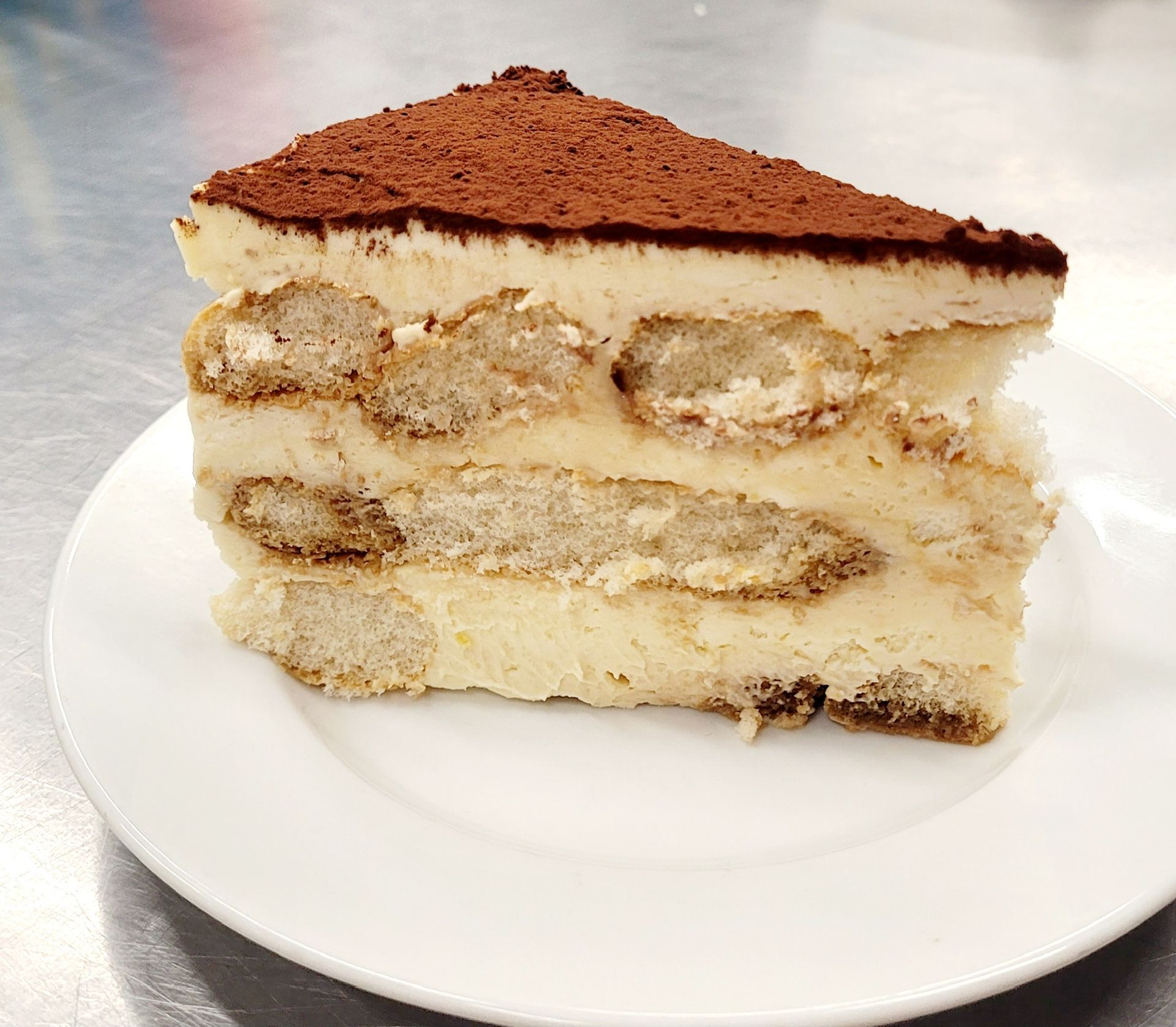 Slice of tiramisu on a white plate, with layers of ladyfingers, cream, and cocoa powder.