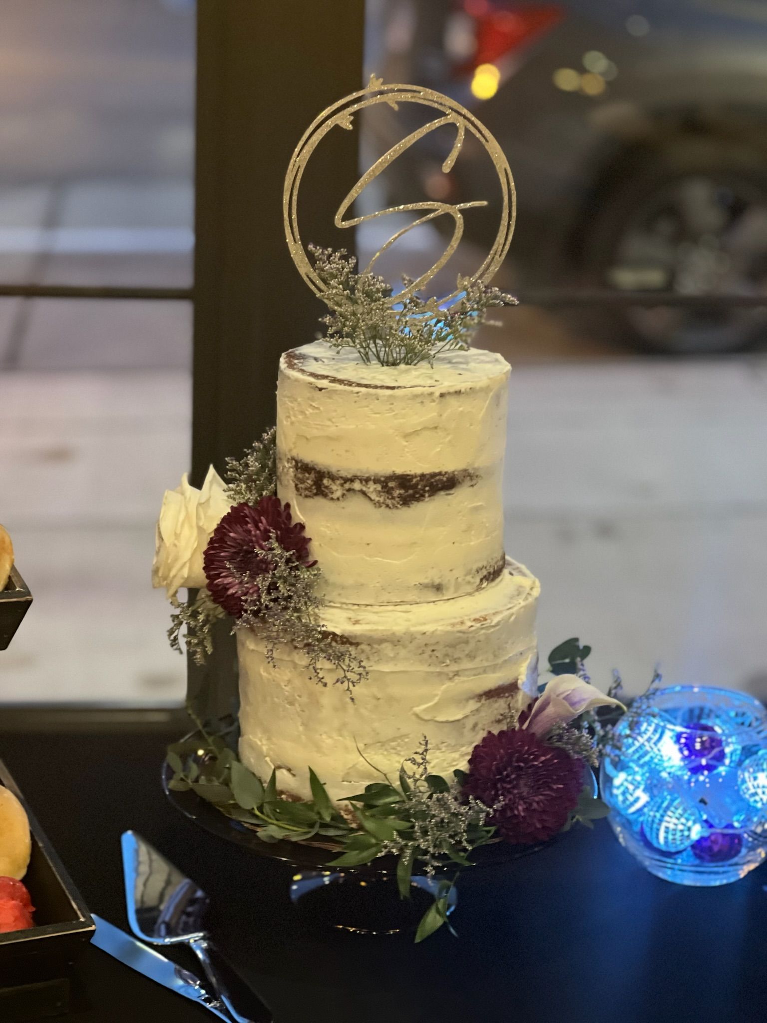 Two-tiered cake with white frosting, floral accents, and a gold monogram topper on a table with a decorative light.