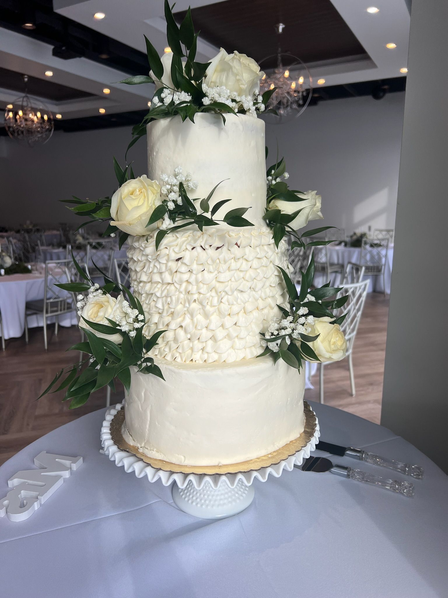 Three-tiered white wedding cake decorated with flowers and greenery on a table in a reception hall.
