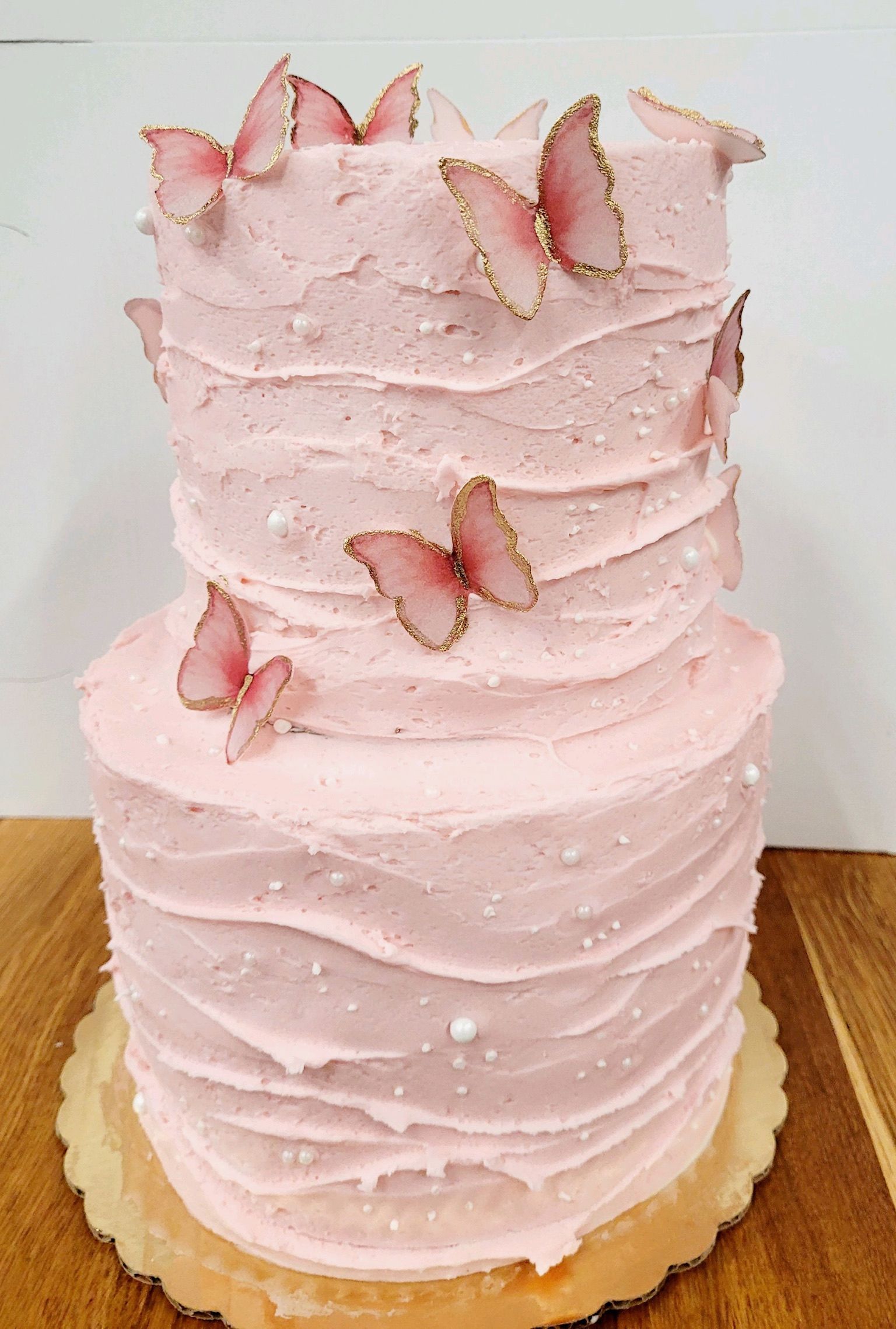Two-tiered pink frosted cake with edible butterfly decorations and a gold base on a wooden table.