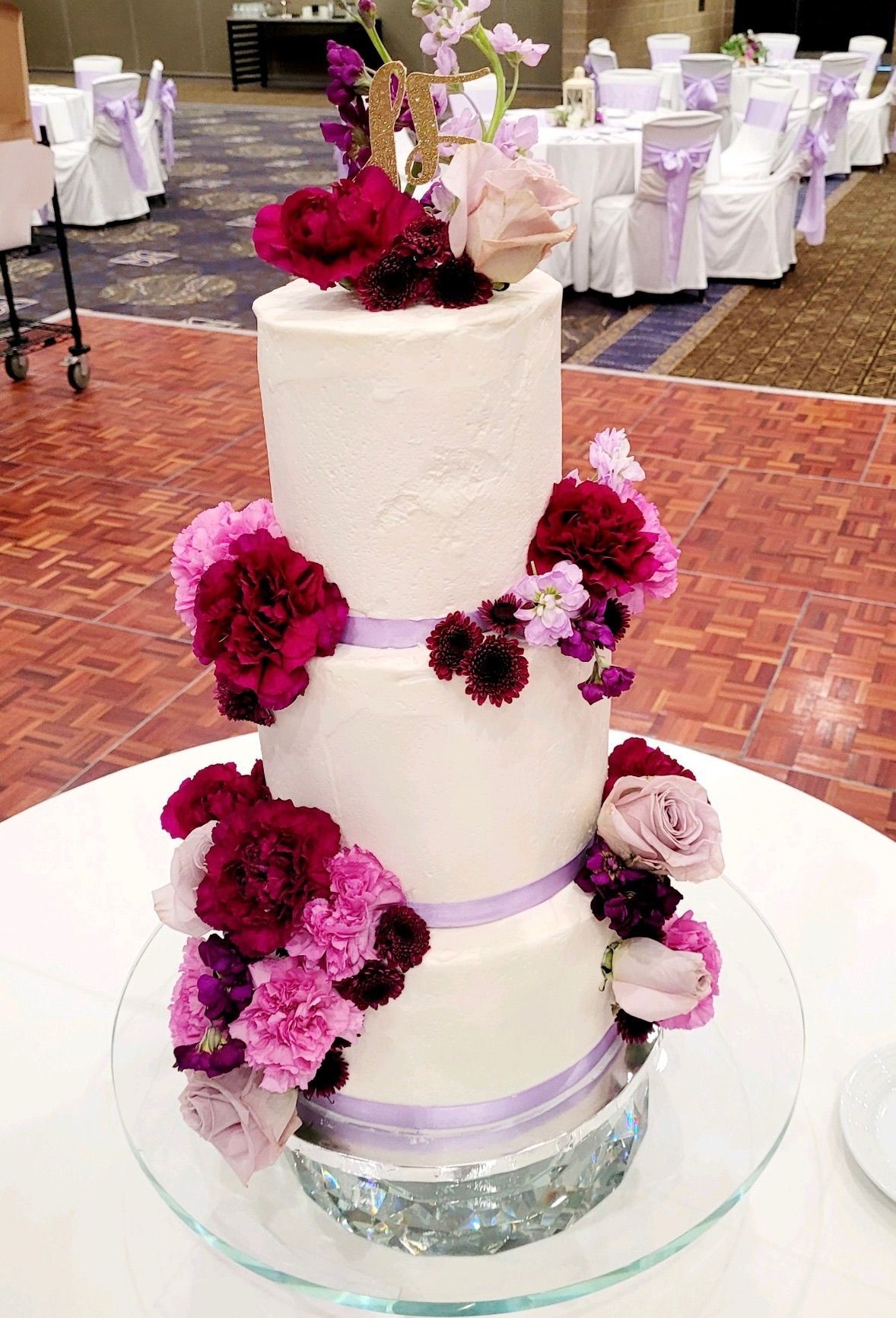 Three-tiered wedding cake, decorated with flowers and ribbons.  Purple, pink, and burgundy flowers adorn the white frosted cake.