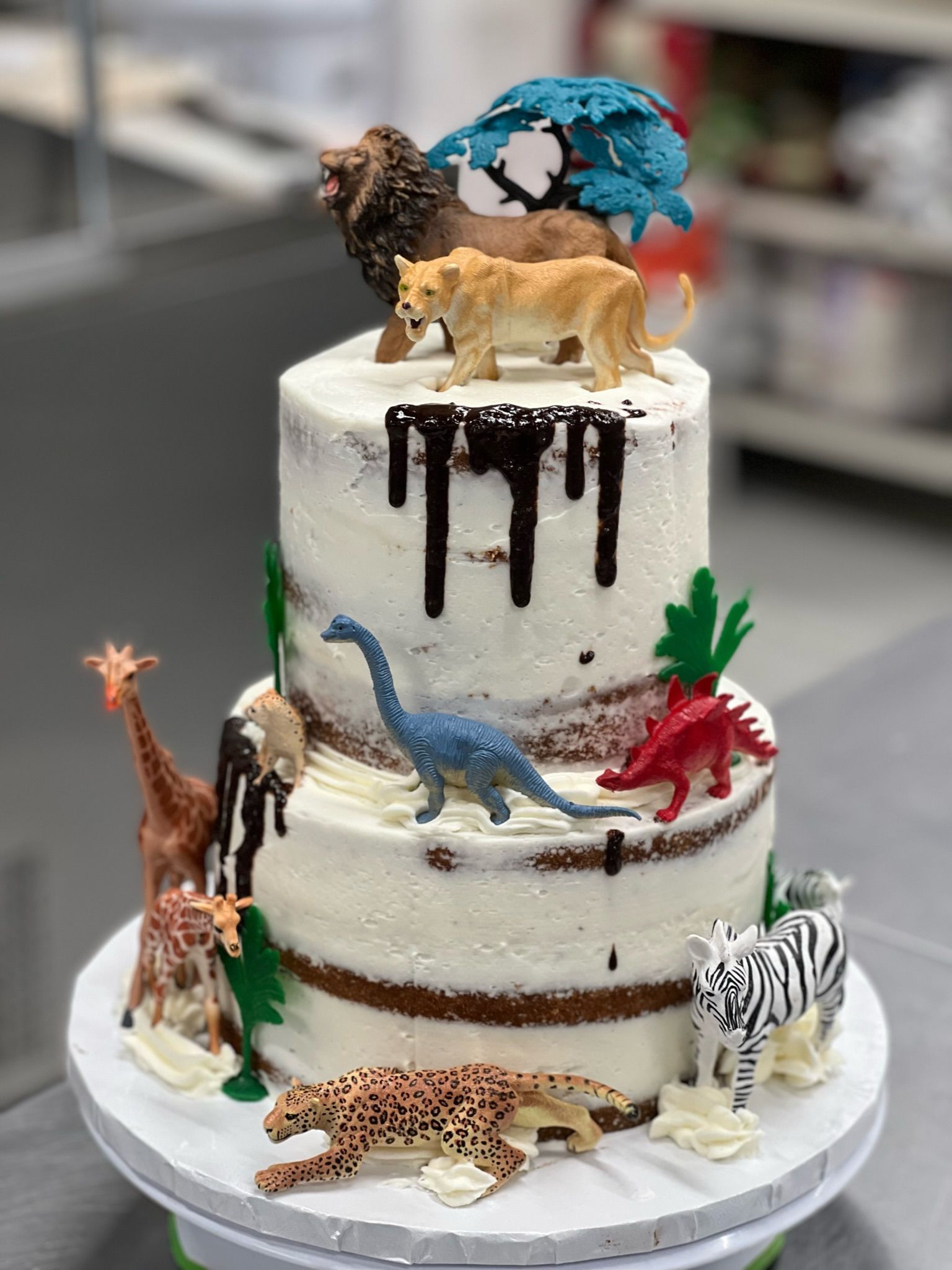 Two-tiered cake decorated with toy animals, chocolate drips, and green accents.