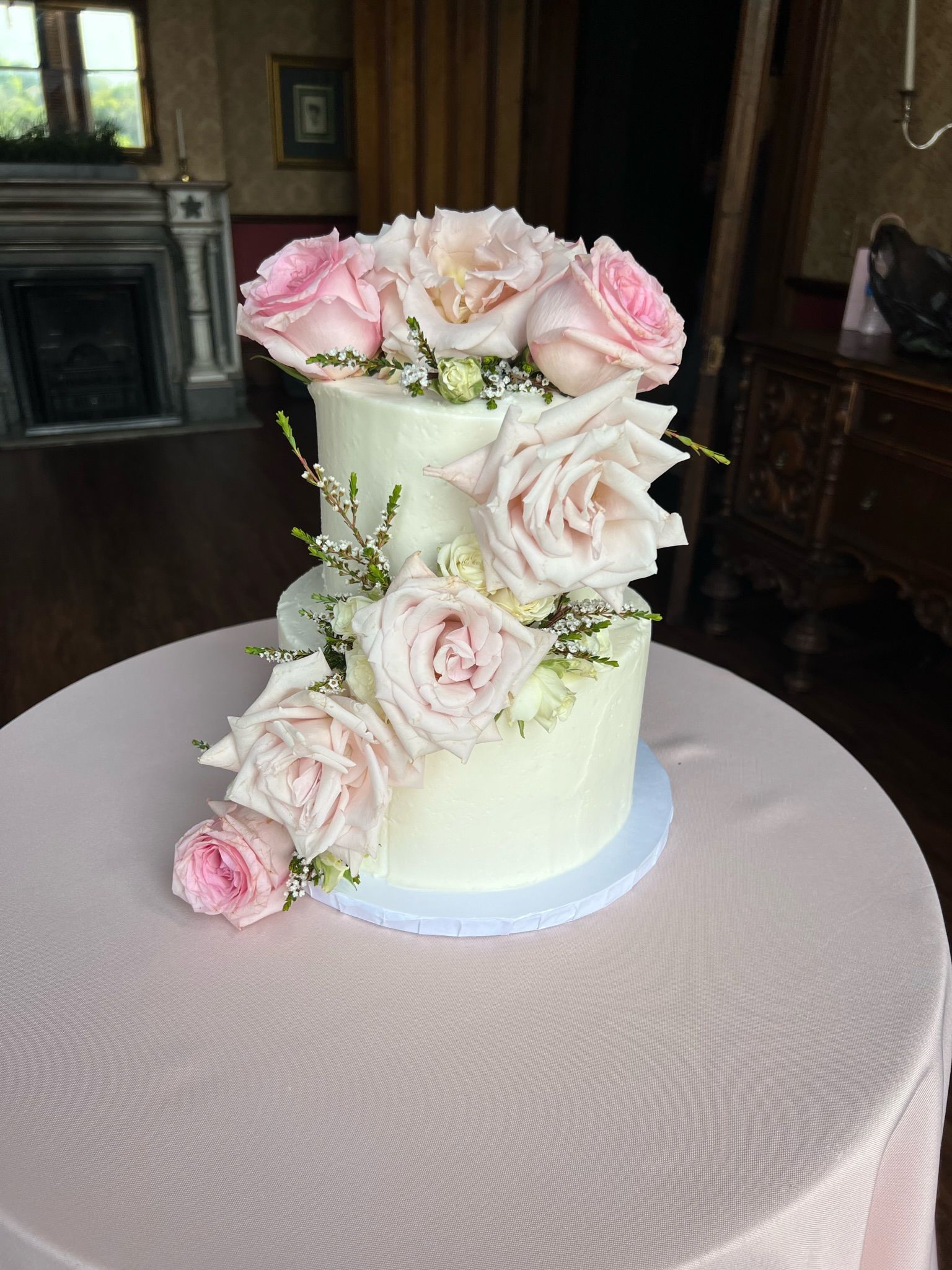 Two-tiered white frosted cake adorned with pink roses, set on a pink tablecloth, in a room with a fireplace.