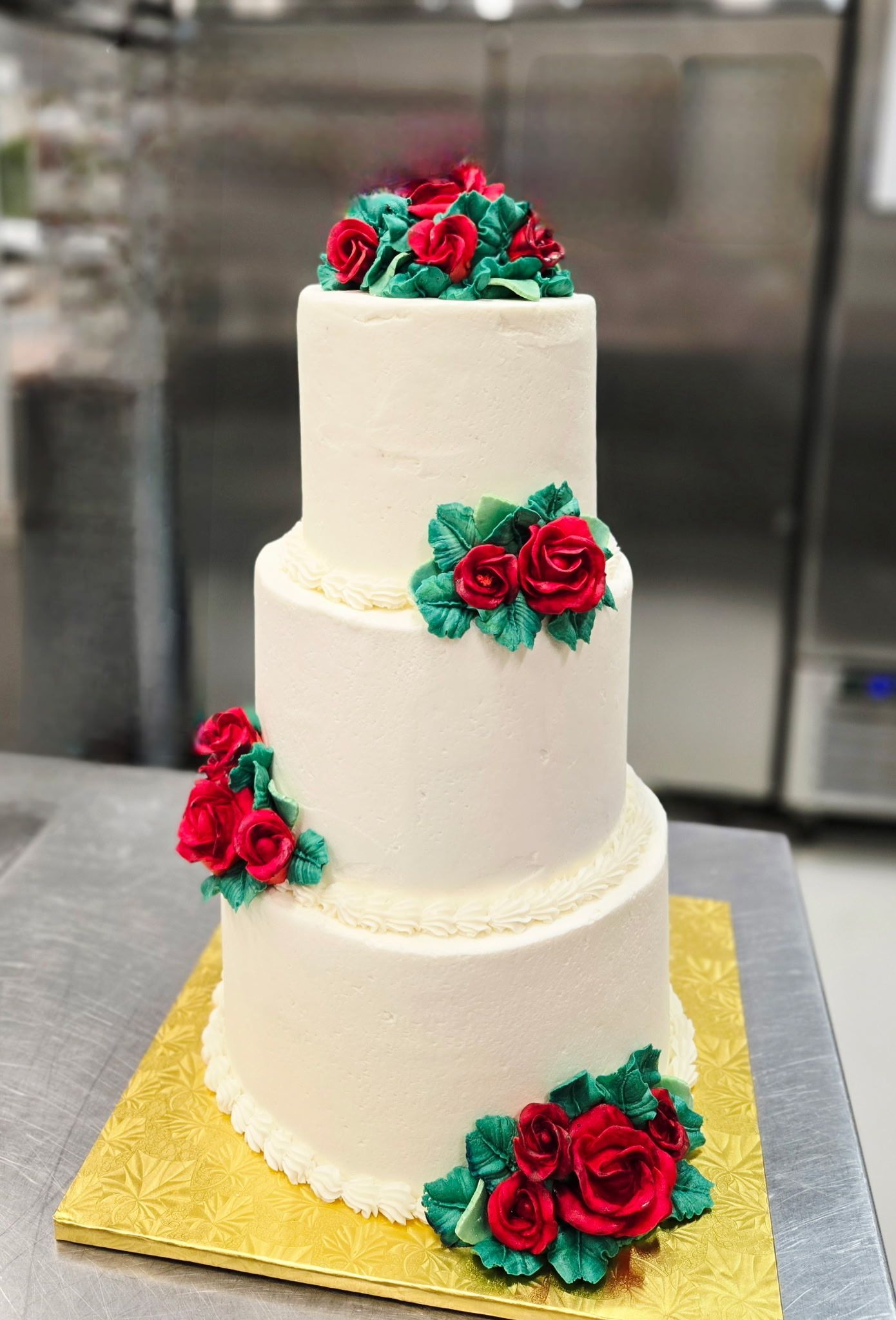 Three-tiered white cake with red rose and green leaf decorations. Gold base, neutral setting.