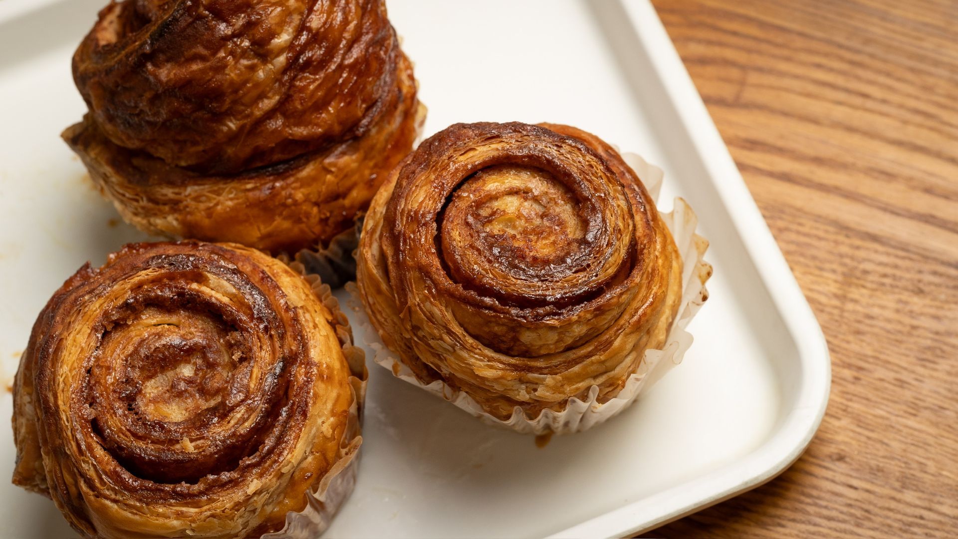 Cinnamon rolls on a white tray, set on a wooden surface. Three rolls are in focus, golden brown and swirled.