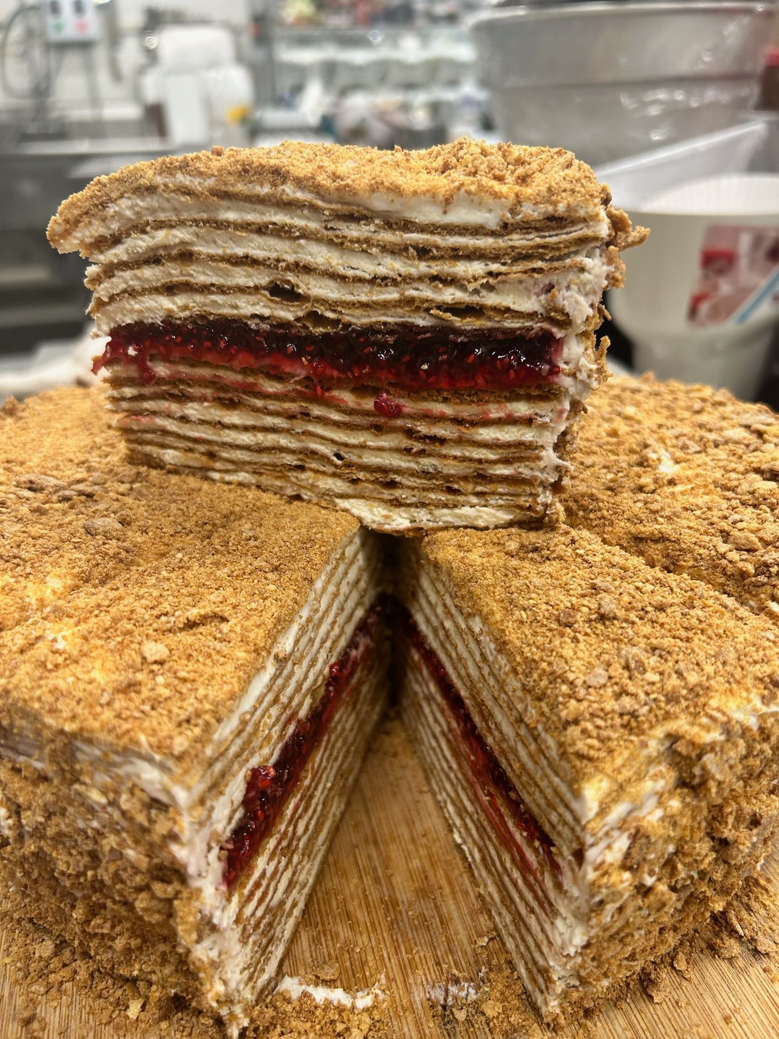 Layered honey cake with red jam filling, topped with crumbs. A slice is removed.