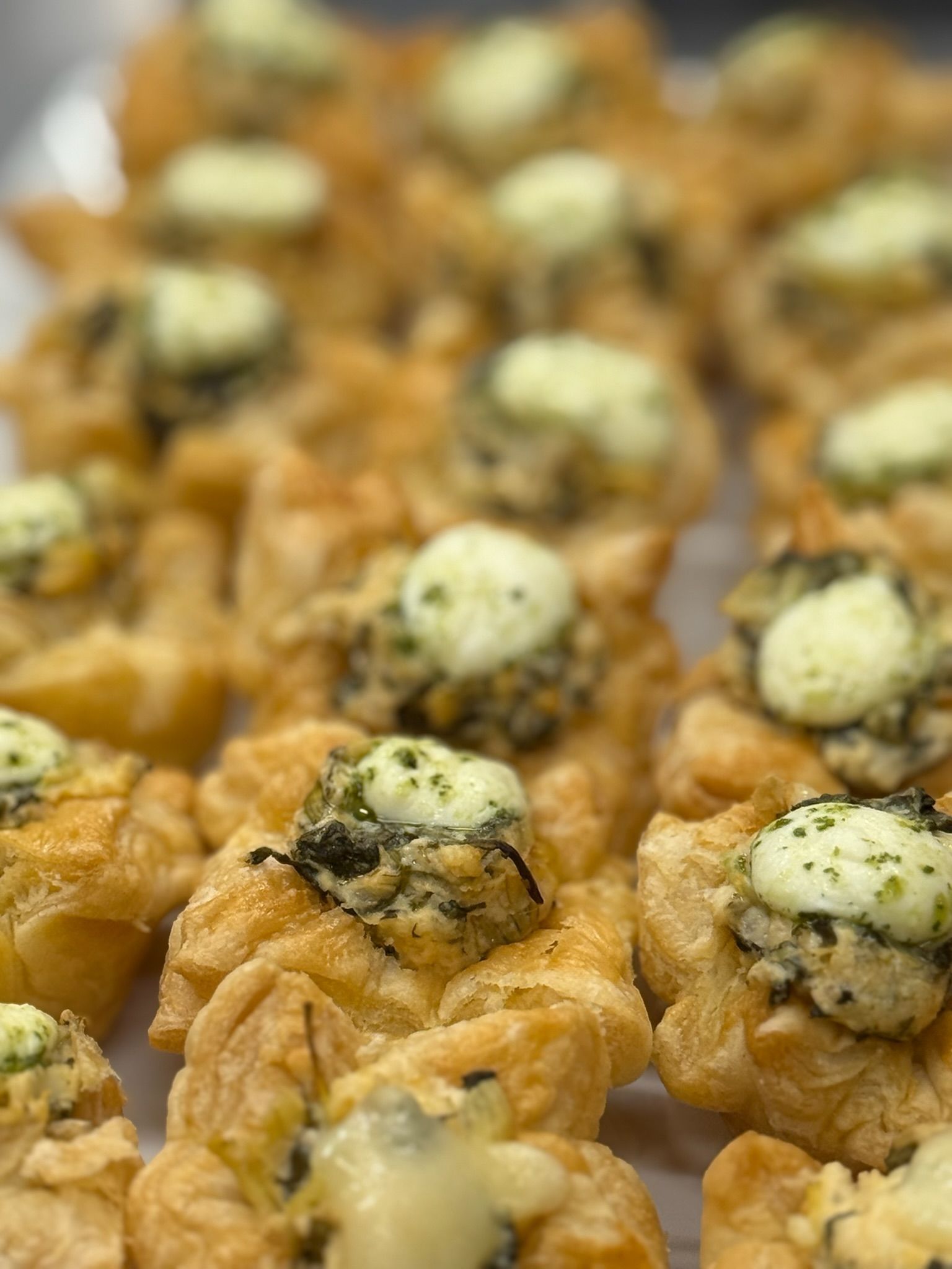Close-up of golden, flaky pastry bites, topped with green herb filling and white cheese.