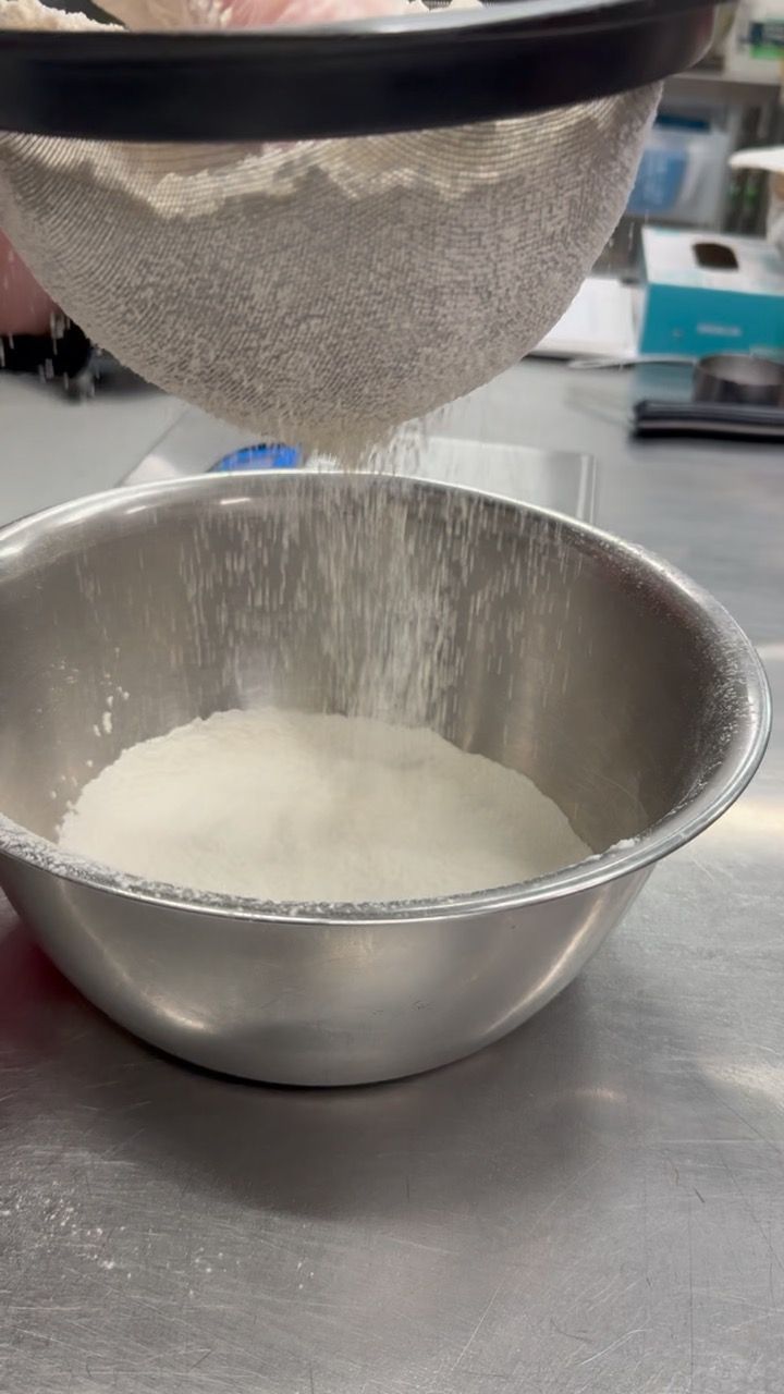 Flour being sifted from a mesh sieve into a stainless steel bowl on a metal surface.