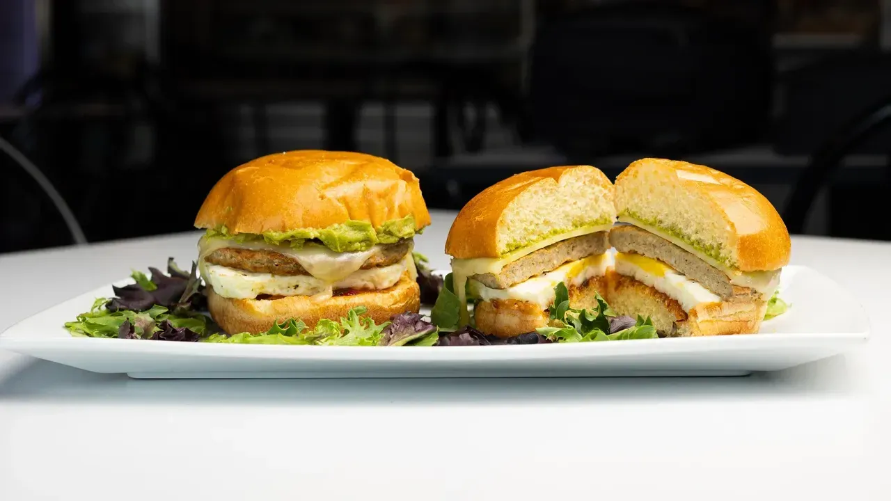 Two halves of a burger on a plate with avocado, cheese, a fried egg and pesto.