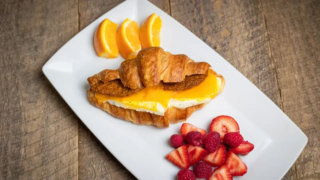 Croissant breakfast sandwich with egg and cheese, orange slices, and berries on a white plate.