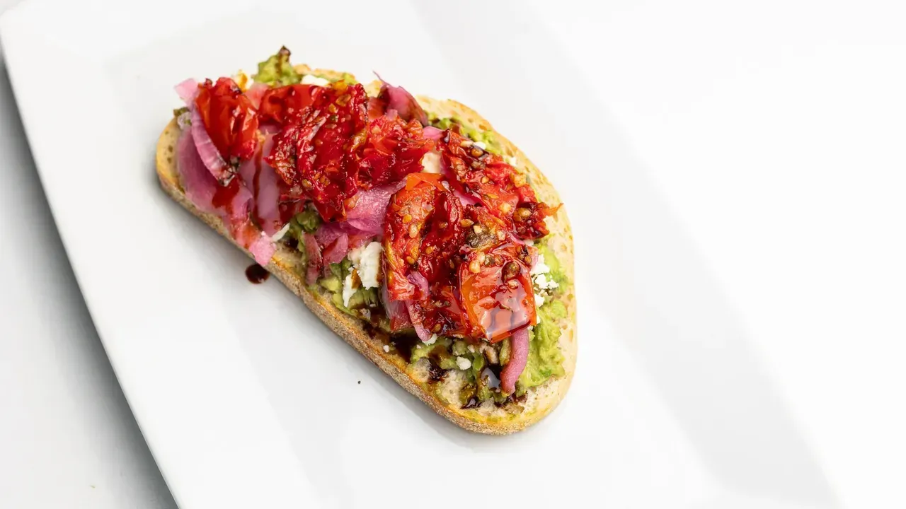 Toasted bread topped with pesto, red onion, sun-dried tomatoes, and a balsamic glaze on a white plate.