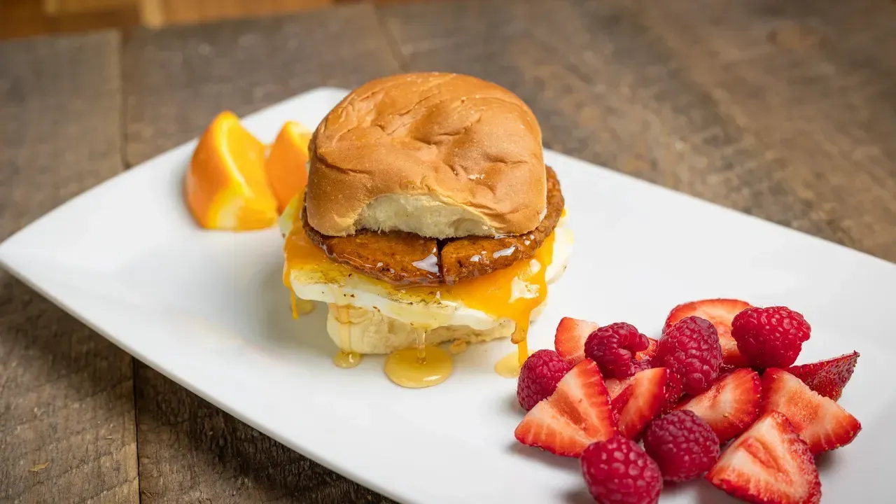 Breakfast sandwich with fruit on a white plate: egg, sausage, cheese, bun, orange slices, strawberries, and raspberries.