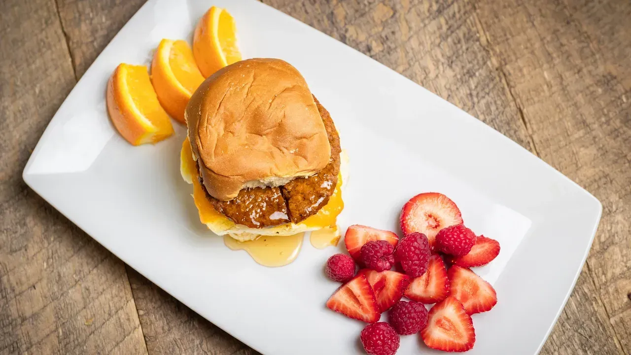 Breakfast sandwich, orange slices, and berries on a white plate atop a wooden surface.