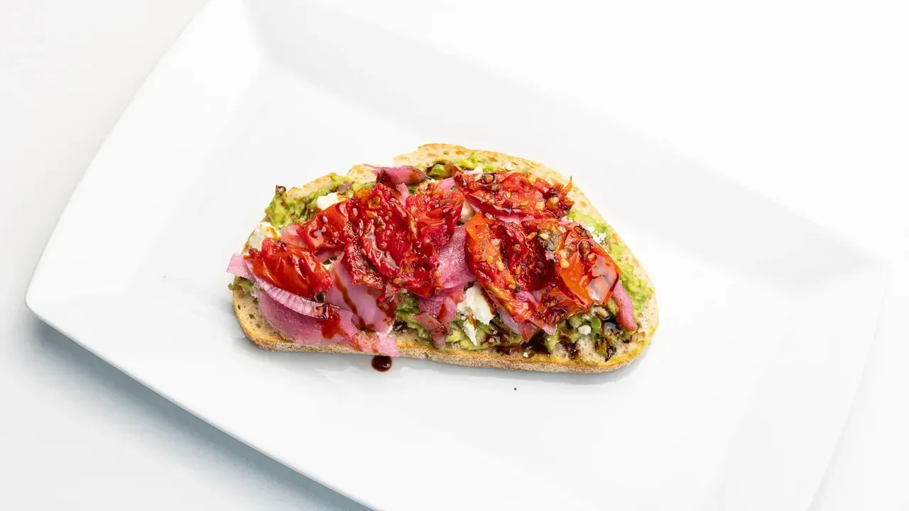 Toast topped with pesto, roasted tomatoes, pickled onions, and balsamic glaze on a white plate.