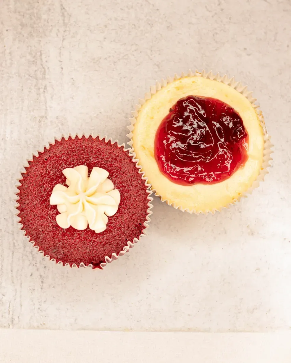 Red velvet cupcake with white frosting and a cheesecake cupcake with red jam on a gray surface.