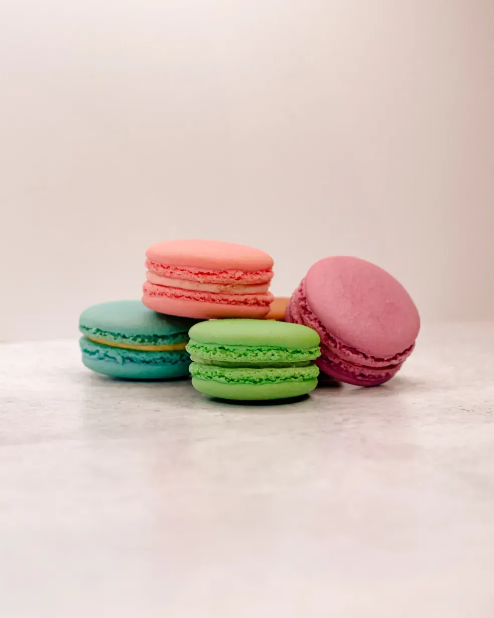 Pile of colorful macarons: pink, green, teal, and purple. On a light-colored surface.