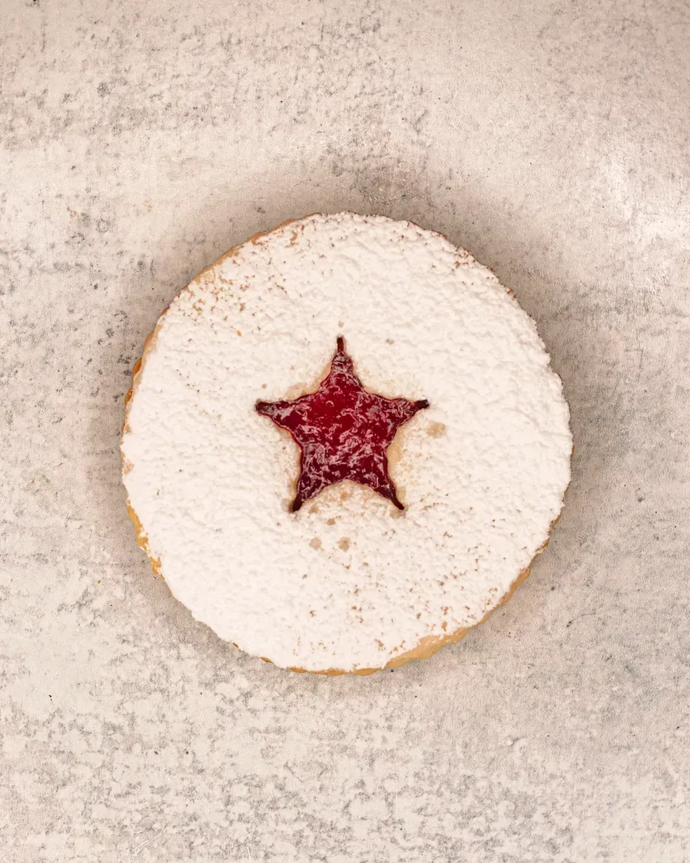 Round cookie with a star-shaped cutout filled with red jam, dusted with powdered sugar.