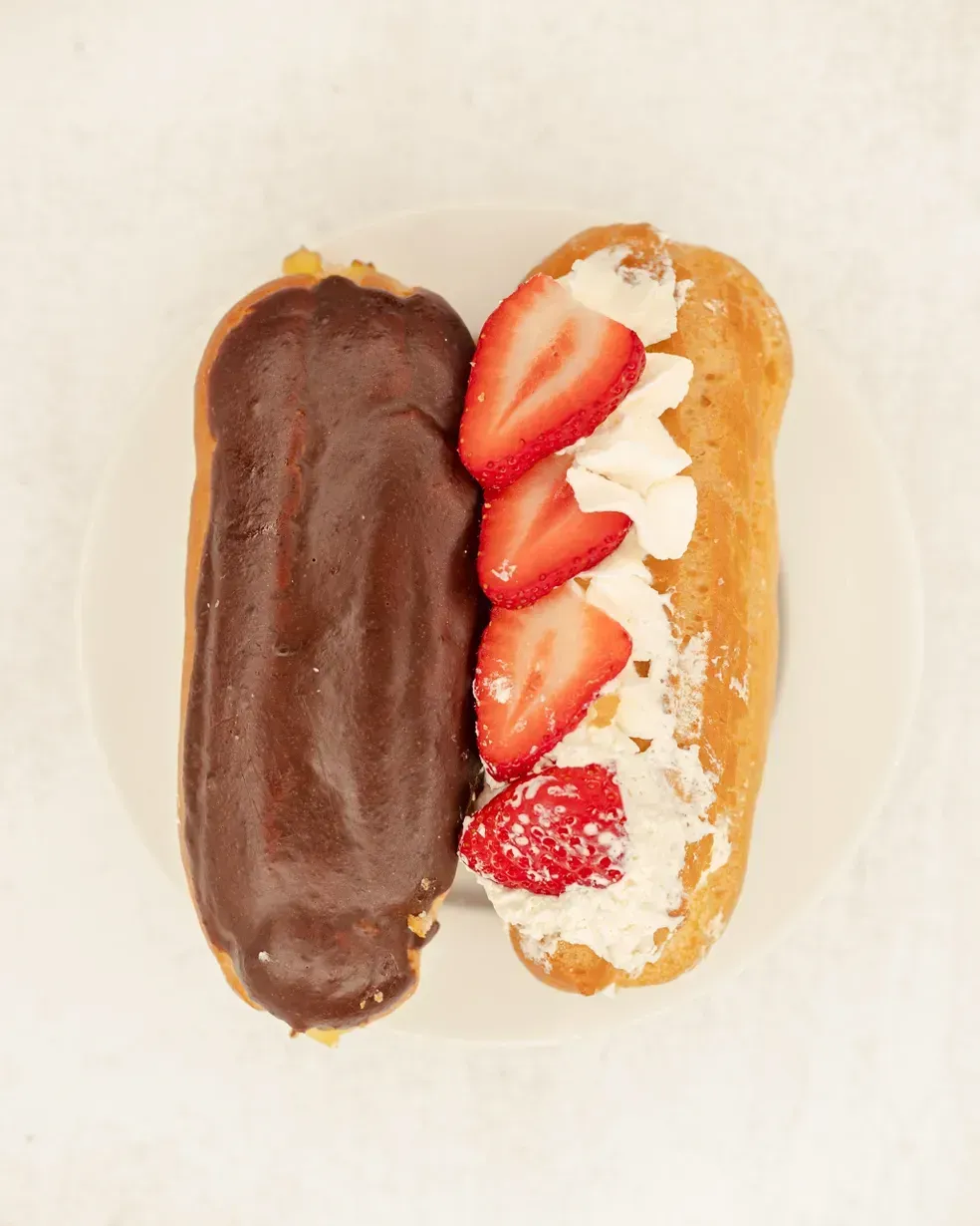 Two eclairs, one chocolate-glazed, one with whipped cream and strawberries, on a white plate.