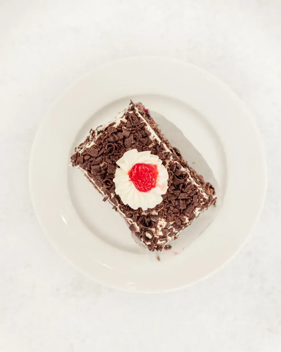 Slice of Black Forest cake on a white plate, topped with whipped cream and a cherry.