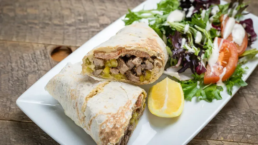 Shawarma wrap cut in half on a plate with greens, tomatoes, and a lemon wedge.