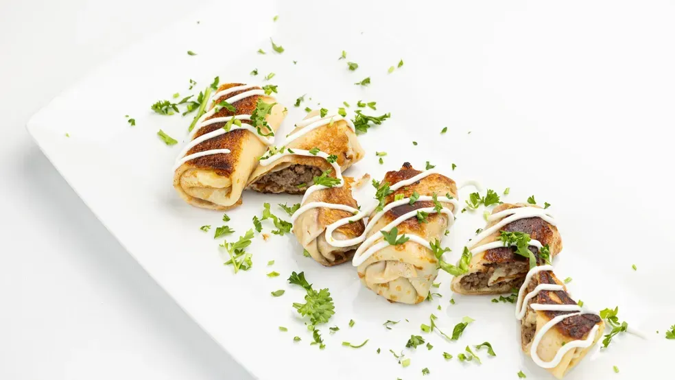 Savory rolled appetizers on a white plate, drizzled with sauce and sprinkled with herbs.