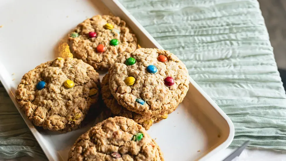 Oatmeal cookies with colorful candies sit on a white tray on a green cloth.