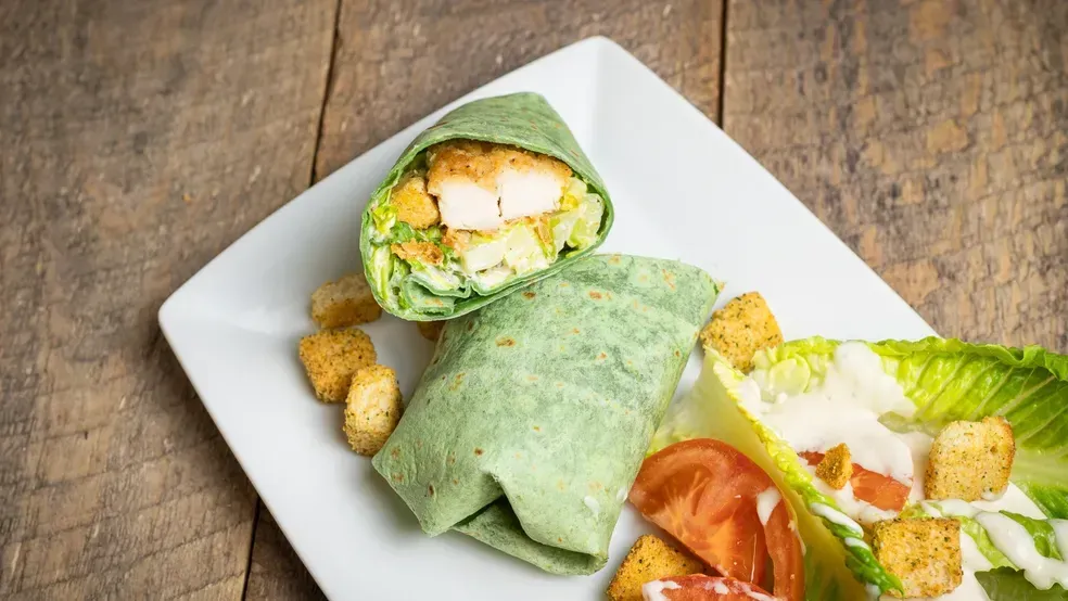 Chicken Caesar wrap on a plate with salad and croutons. Green wrap, wooden table background.