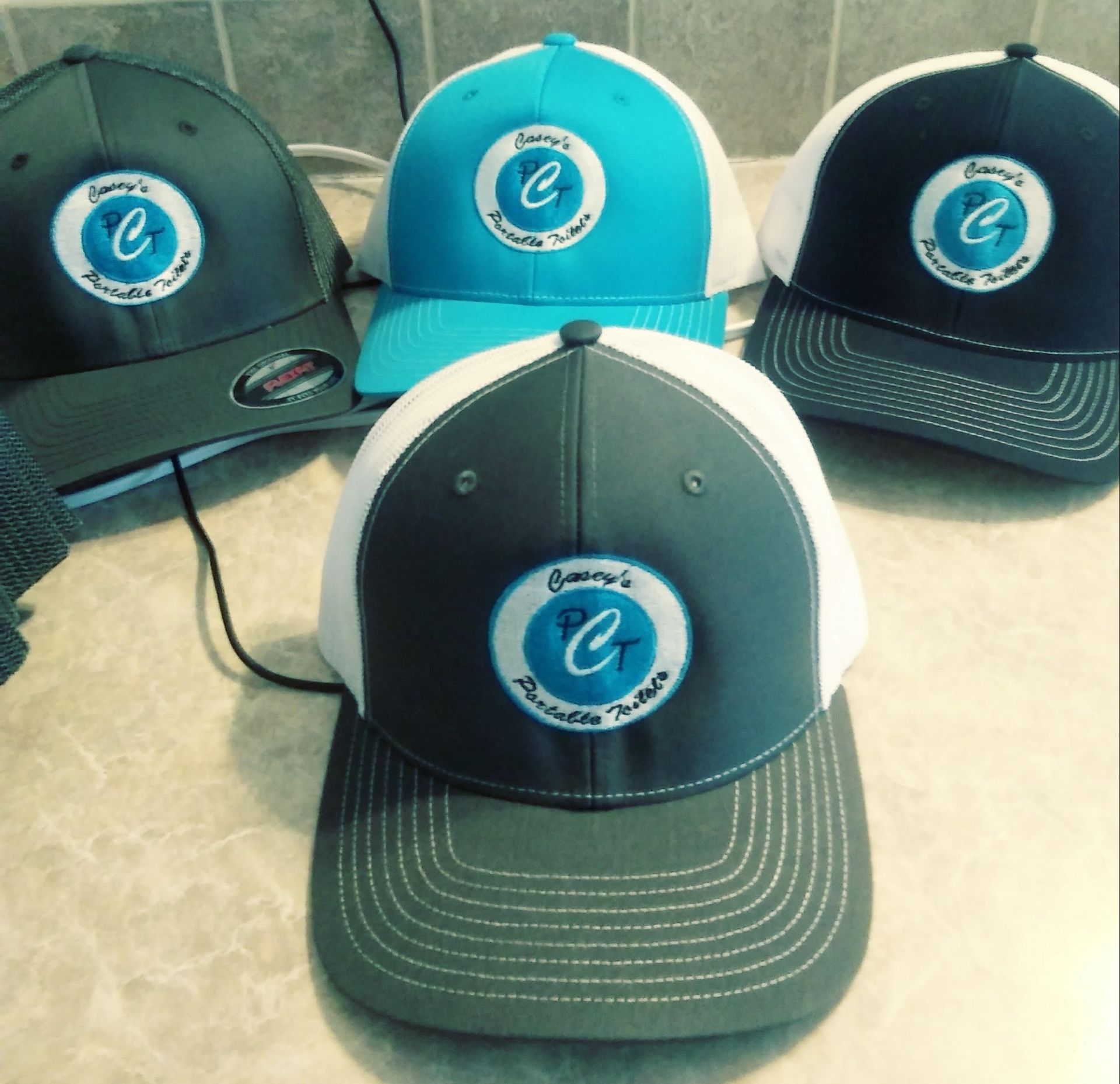 Four baseball caps with custom logo in various colors: gray, turquoise, navy, and charcoal with white mesh back.