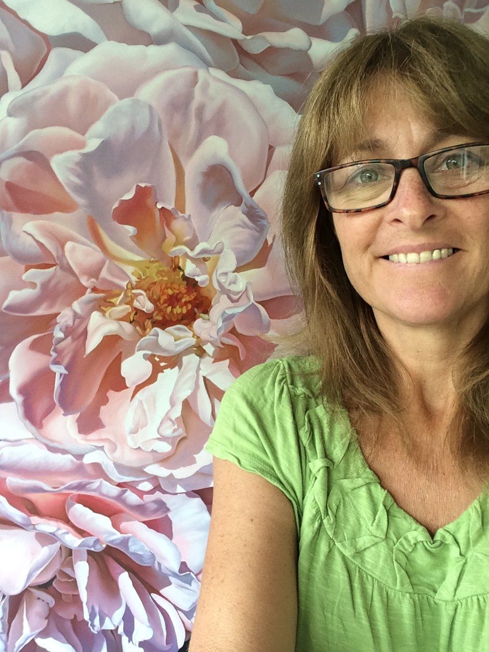 A woman wearing glasses is smiling in front of a painting of a flower.