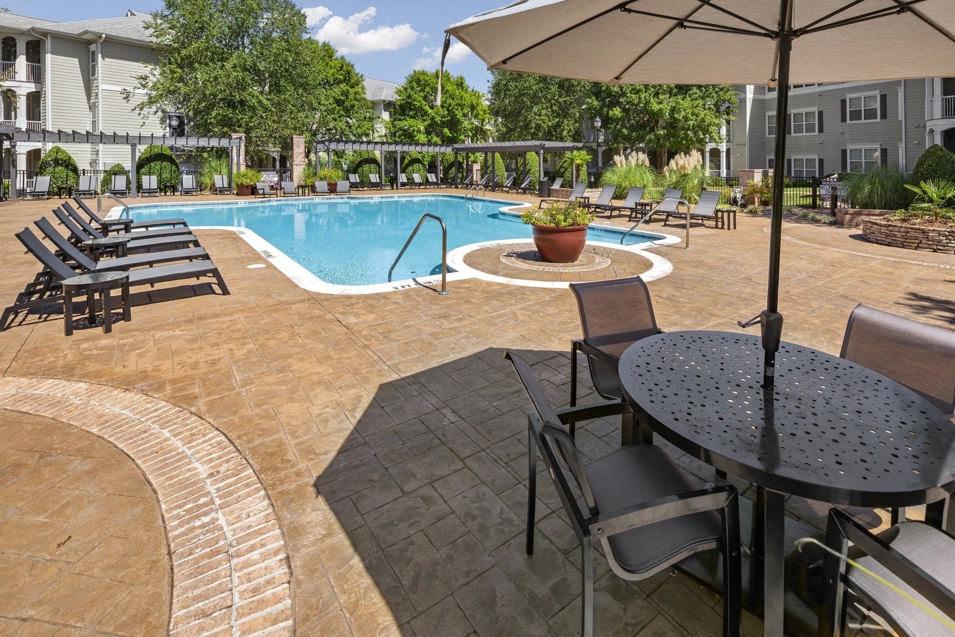 Pool area with lounge chairs, tables, and an umbrella; apartment buildings in background.