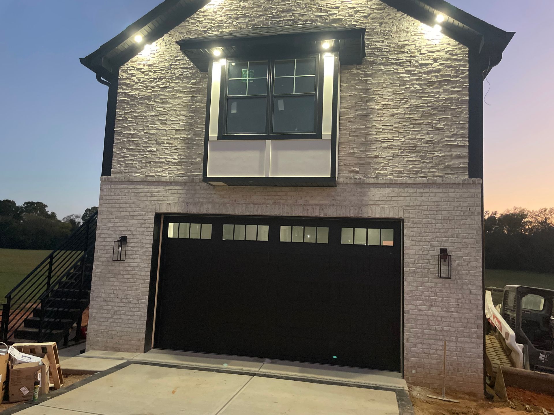 New brick house with black garage door and lit exterior lights at dusk