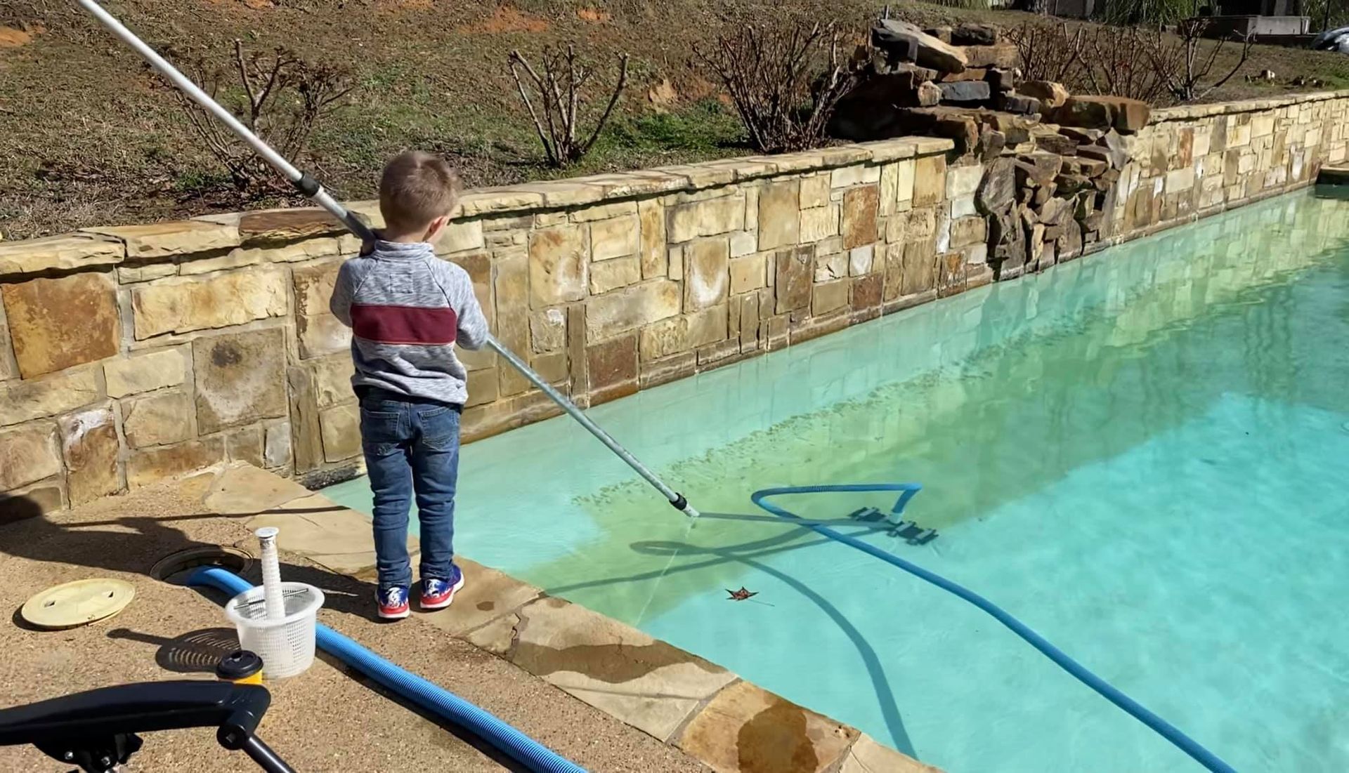 A young boy cleaning a pool with a long-handled net. He wears a striped shirt and jeans near a stone wall.