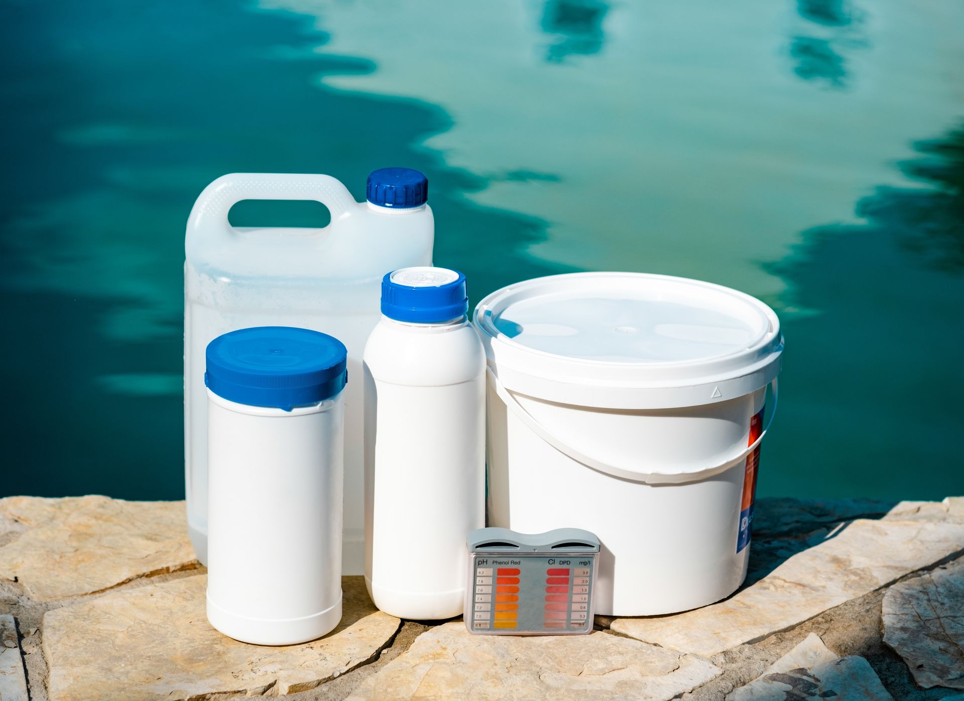 Pool chemicals and test strips on a stone ledge, with a turquoise pool in the background.