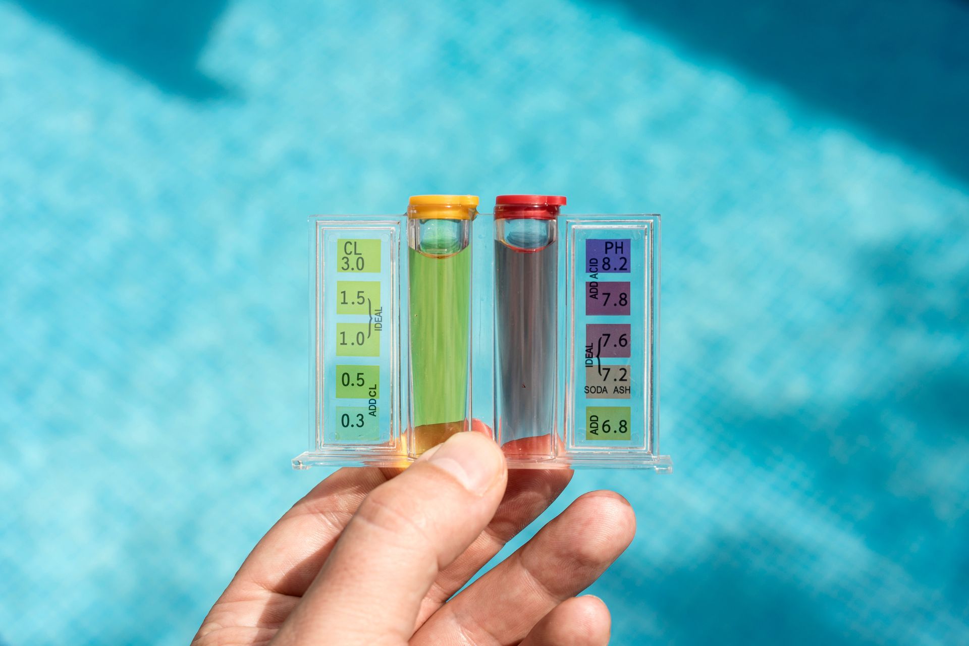 Hand holding a pool water testing kit with green and red tinted water samples against a blue pool.