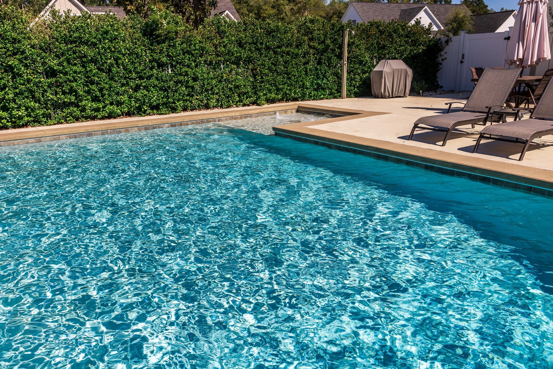 Swimming pool with clear blue water next to a patio with lounge chairs, surrounded by green hedges.