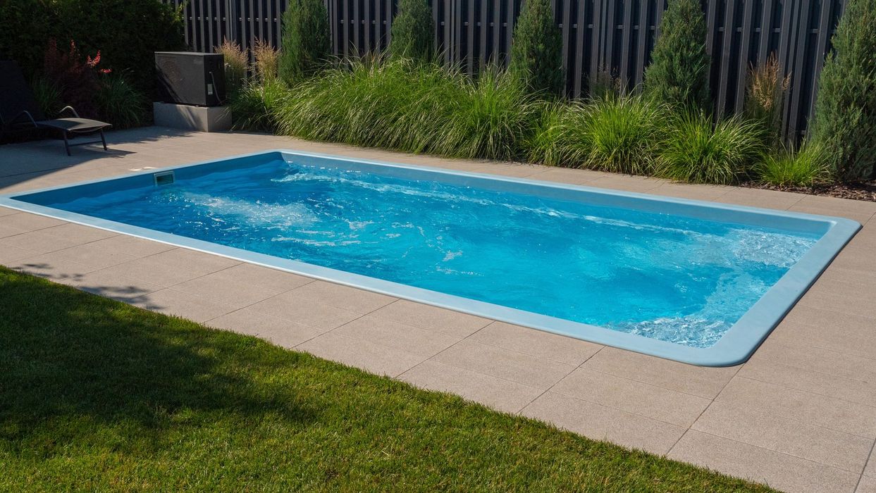 Rectangular blue pool with concrete border, green lawn, and hedge in backyard.