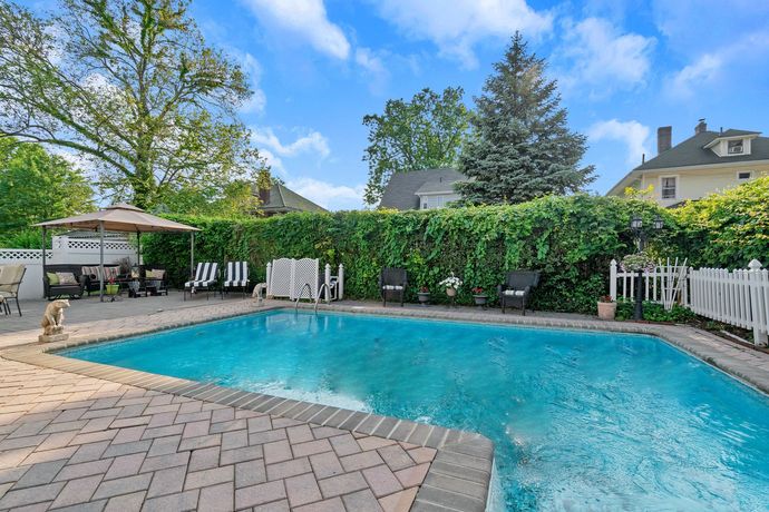 A backyard pool surrounded by brick patio, green foliage, and outdoor seating under a sunny sky.