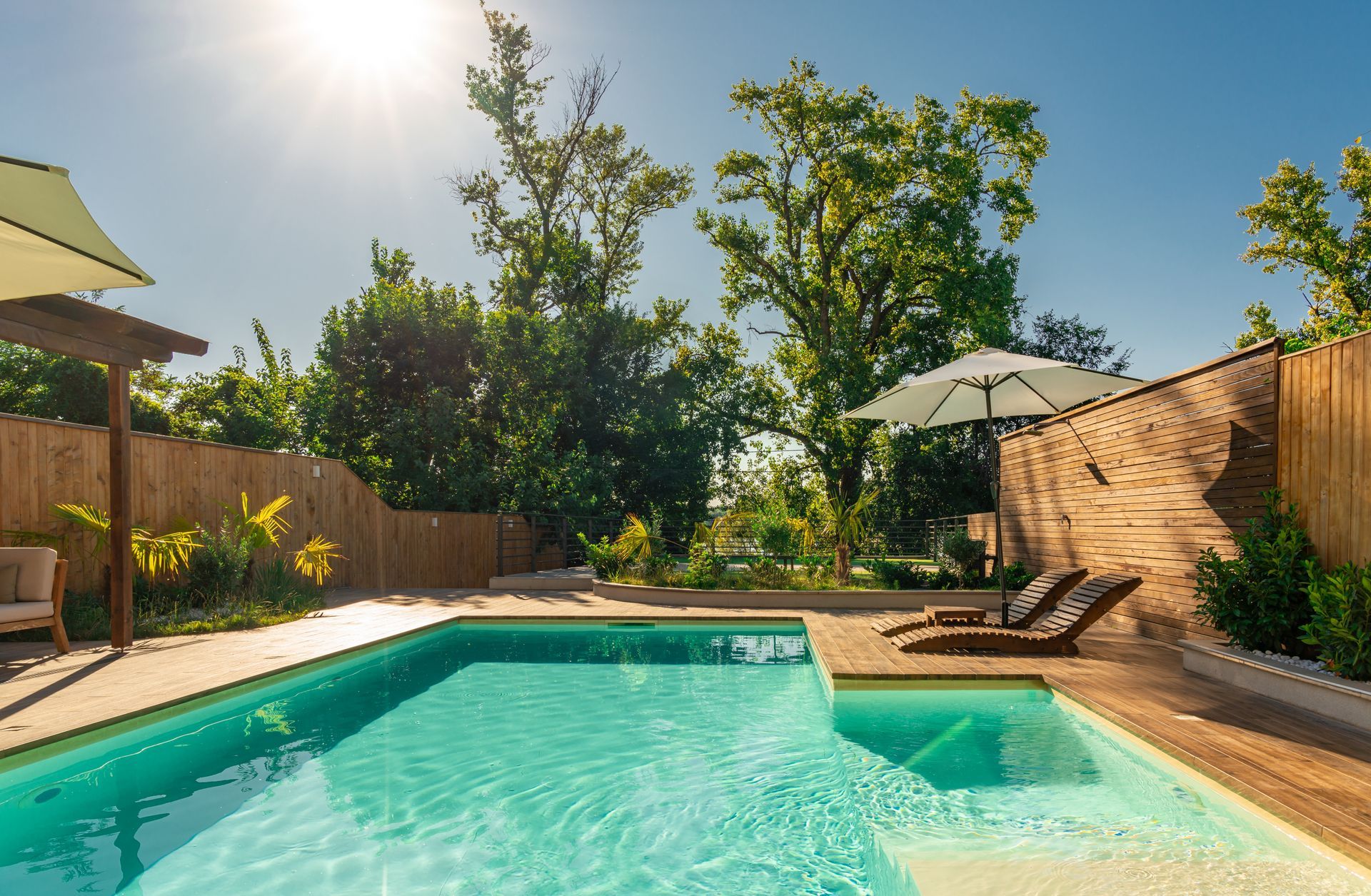 Swimming pool with lounge chairs, blue water, wooden deck, sun shining, trees, and bamboo fence.