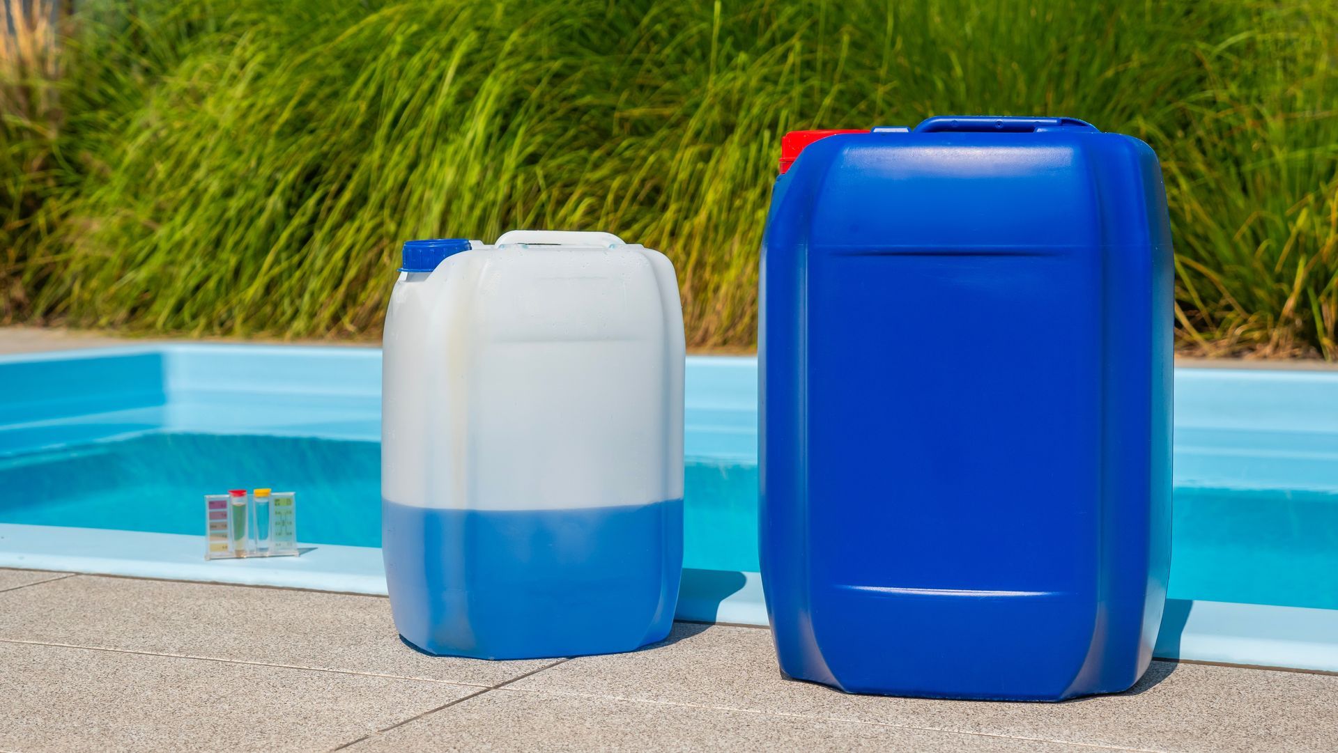 Two blue chemical containers beside a pool with a test kit in the sunlight.