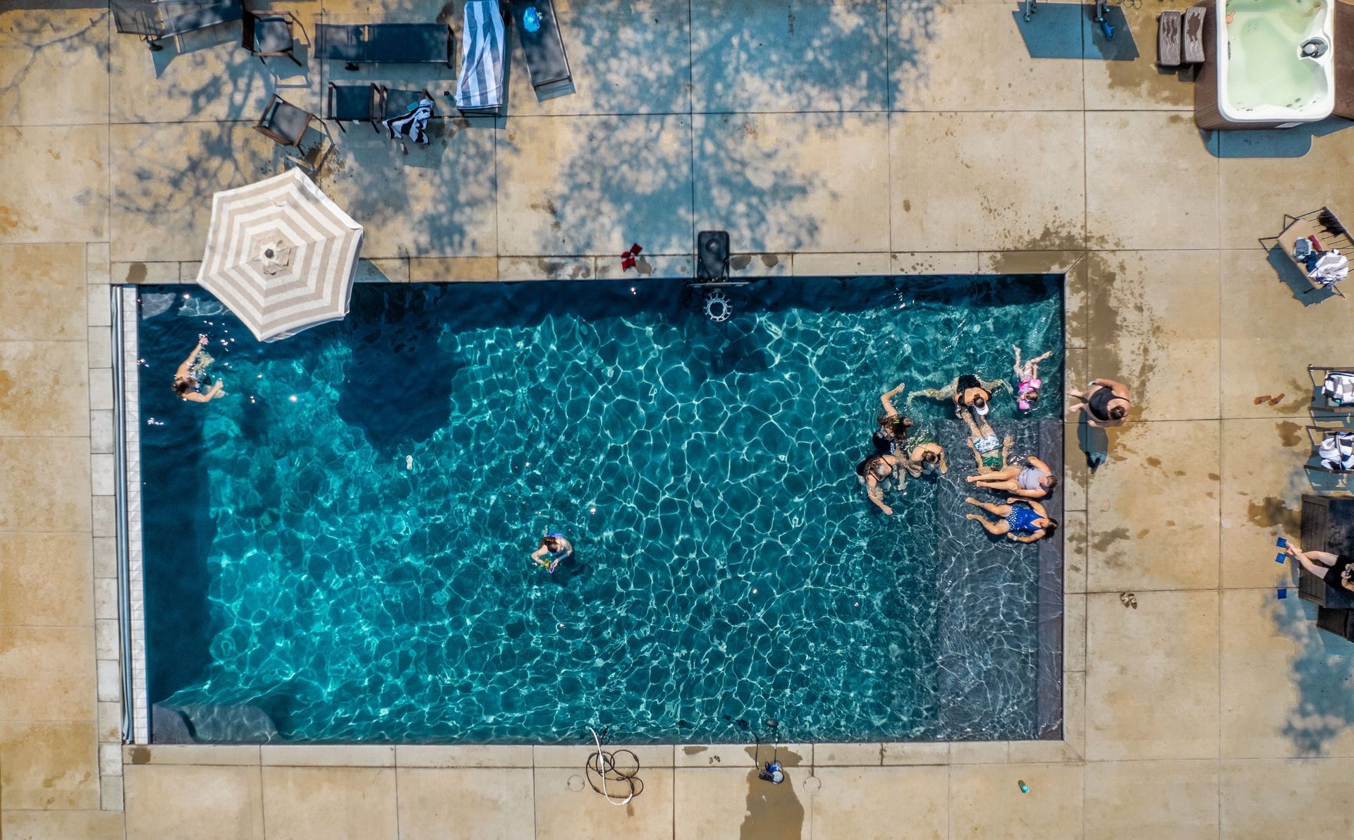 Aerial view of a rectangular pool with people swimming and relaxing. Sun umbrella and surrounding concrete.