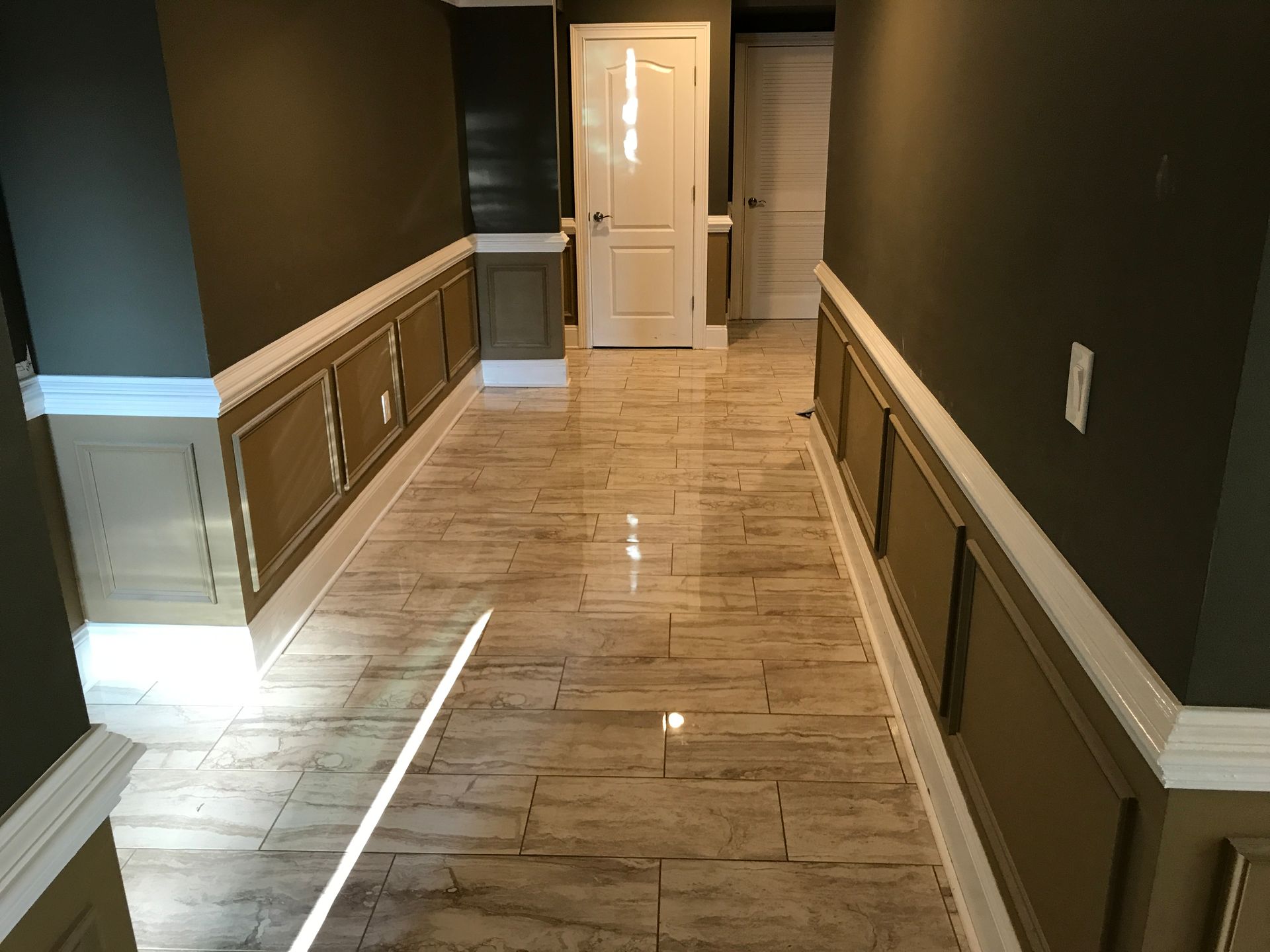 Long hallway with glossy tile floor, dark walls, wainscoting, and a white door at the end