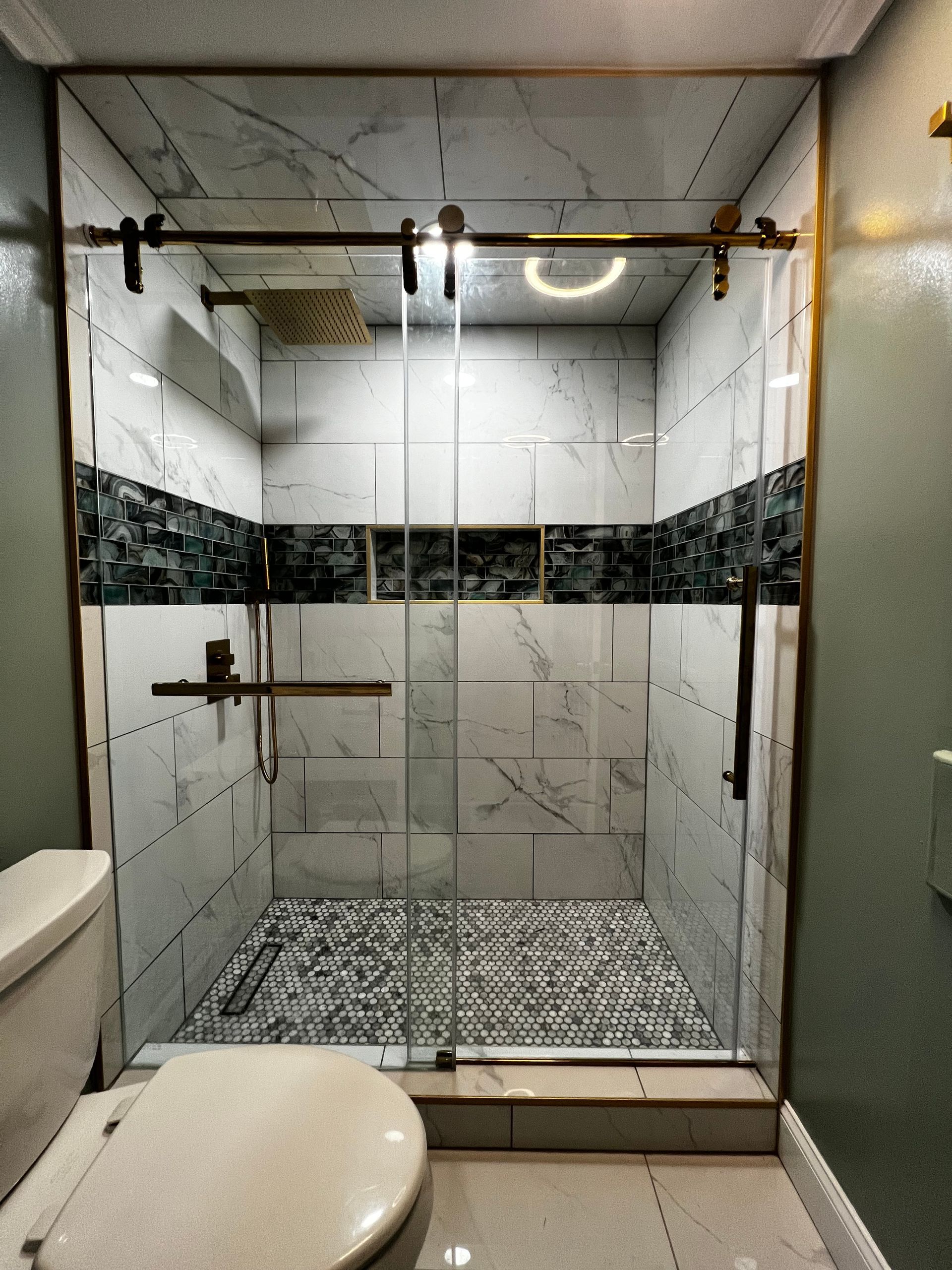 A walk-in shower with white marble-look tiles, a dark mosaic accent strip, gold hardware, and a glass sliding door